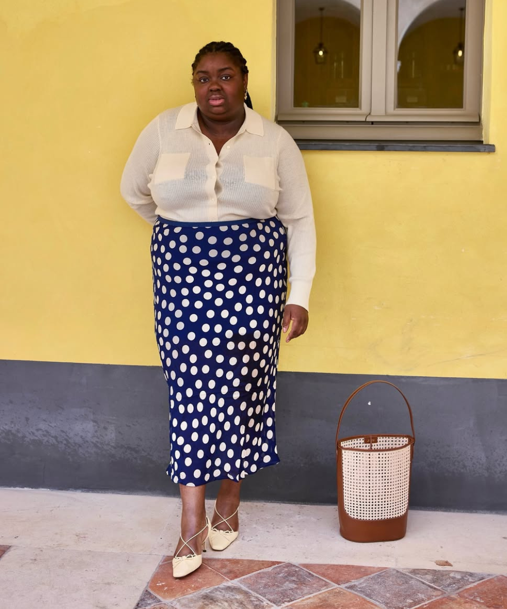 Mulher com saia de poá azul e branca, blusa creme, sapatos claros, em frente a parede amarela.