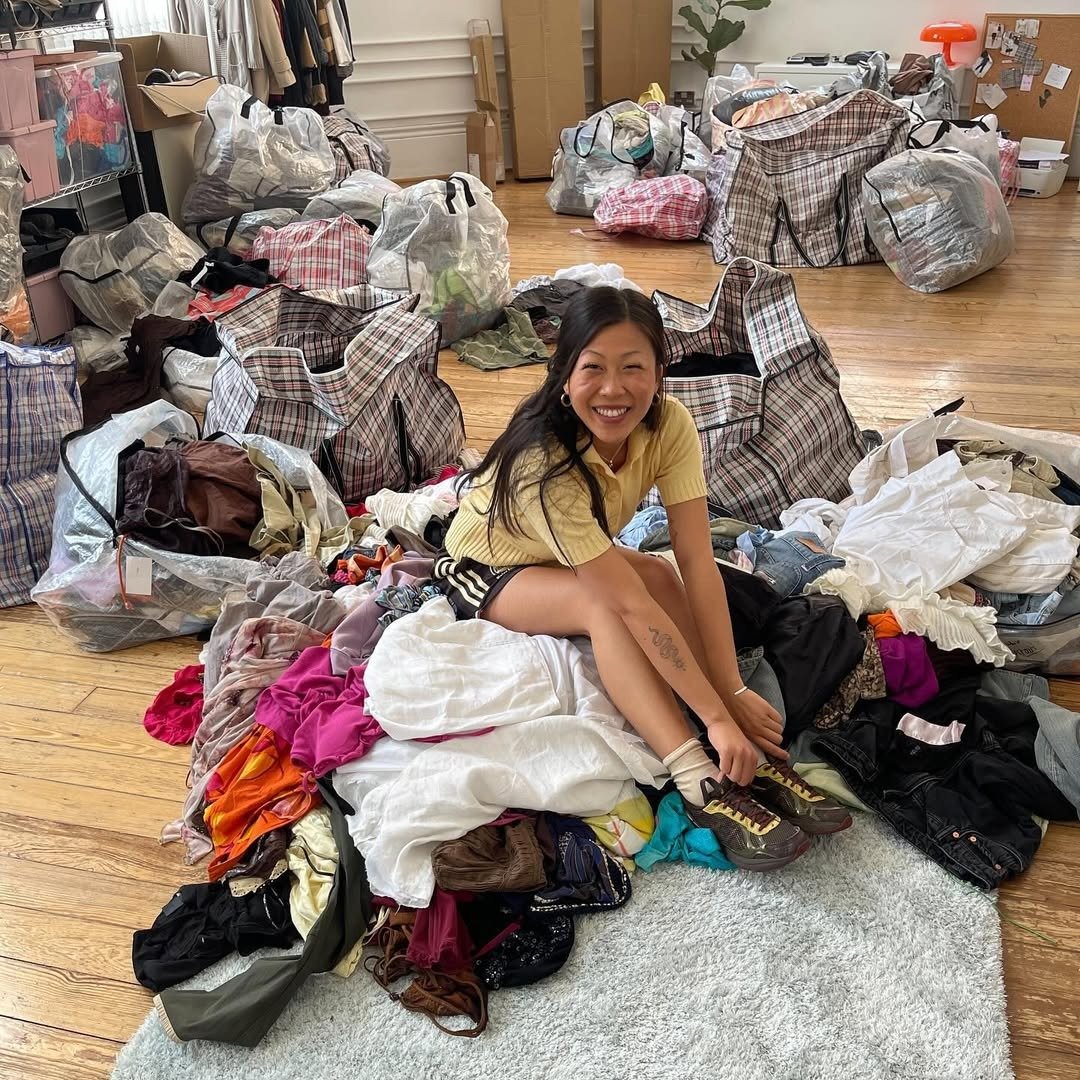 Pessoa sorridente sentada em pilha de roupas em ambiente interno, cercada por bolsas. Ligação entre moda e sustentabilidade.