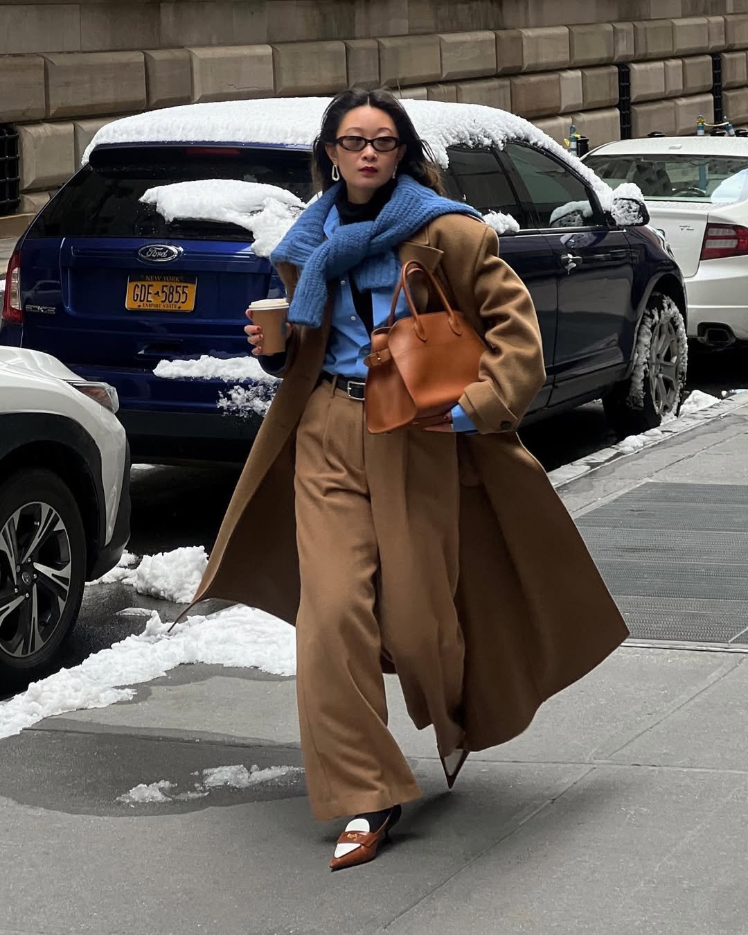 Mulher usa cores que combinam com marrom, como um casaco com tricô azul. Ela está segurando café e bolsa. Fundo de carros e neve.