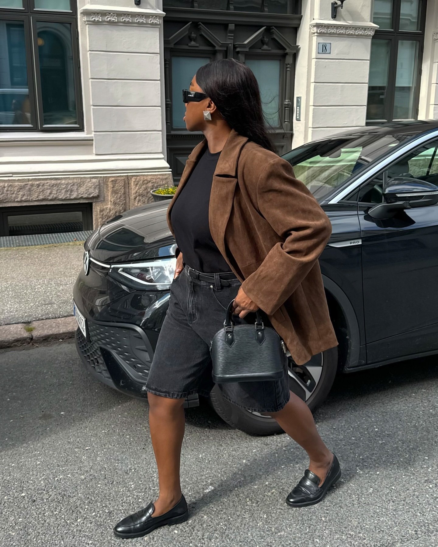 Mulher caminha pela rua, usando blazer marrom que contrasta com roupas pretas e bolsa preta.