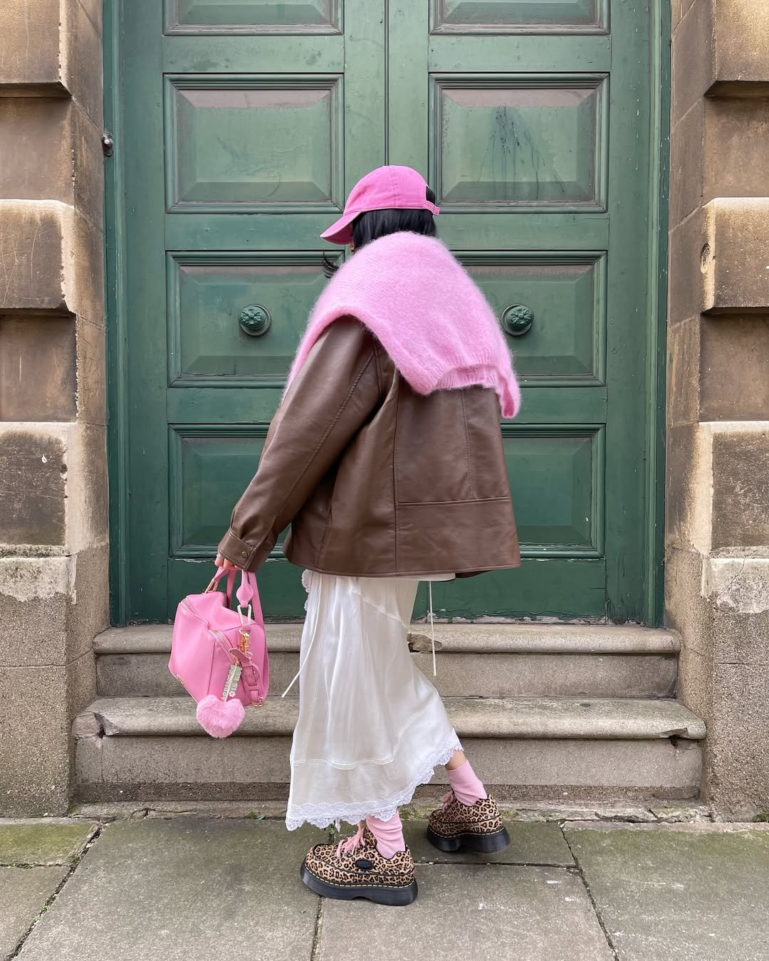 Pessoa de jaqueta marrom, cachecol e bolsa rosa, meias rosa e sapatos de estampa animal. Fundo de porta verde.