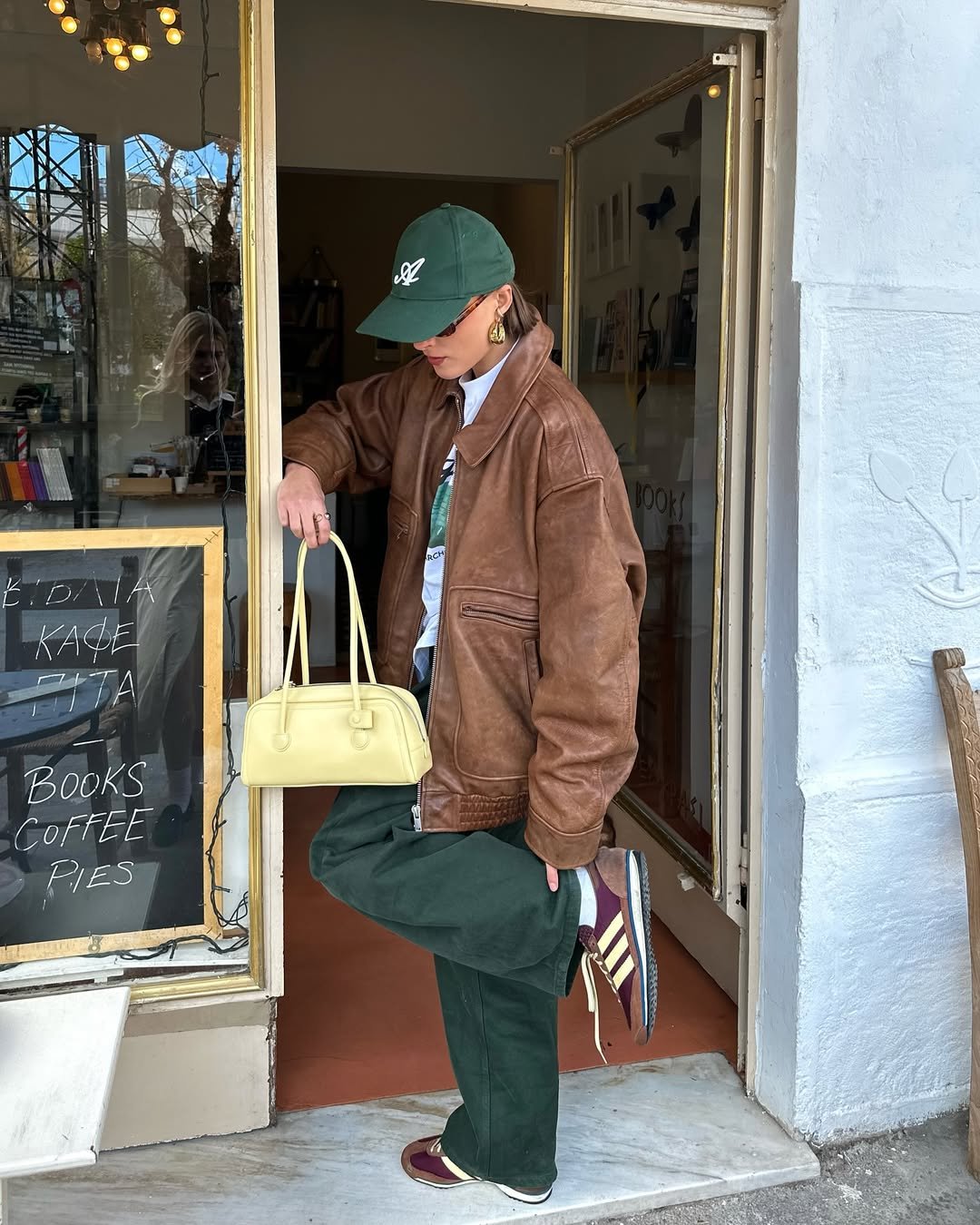 Pessoa com jaqueta marrom, boné verde e bolsa creme, combinando as cores. Em frente a uma loja com livros e café.