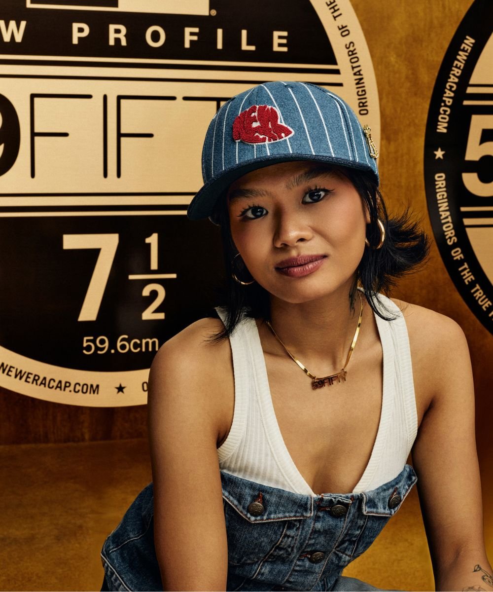 Modelo usa boné da marca 59FIFTY, camisa listrada e colar. Fundo com cartazes e logotipos.