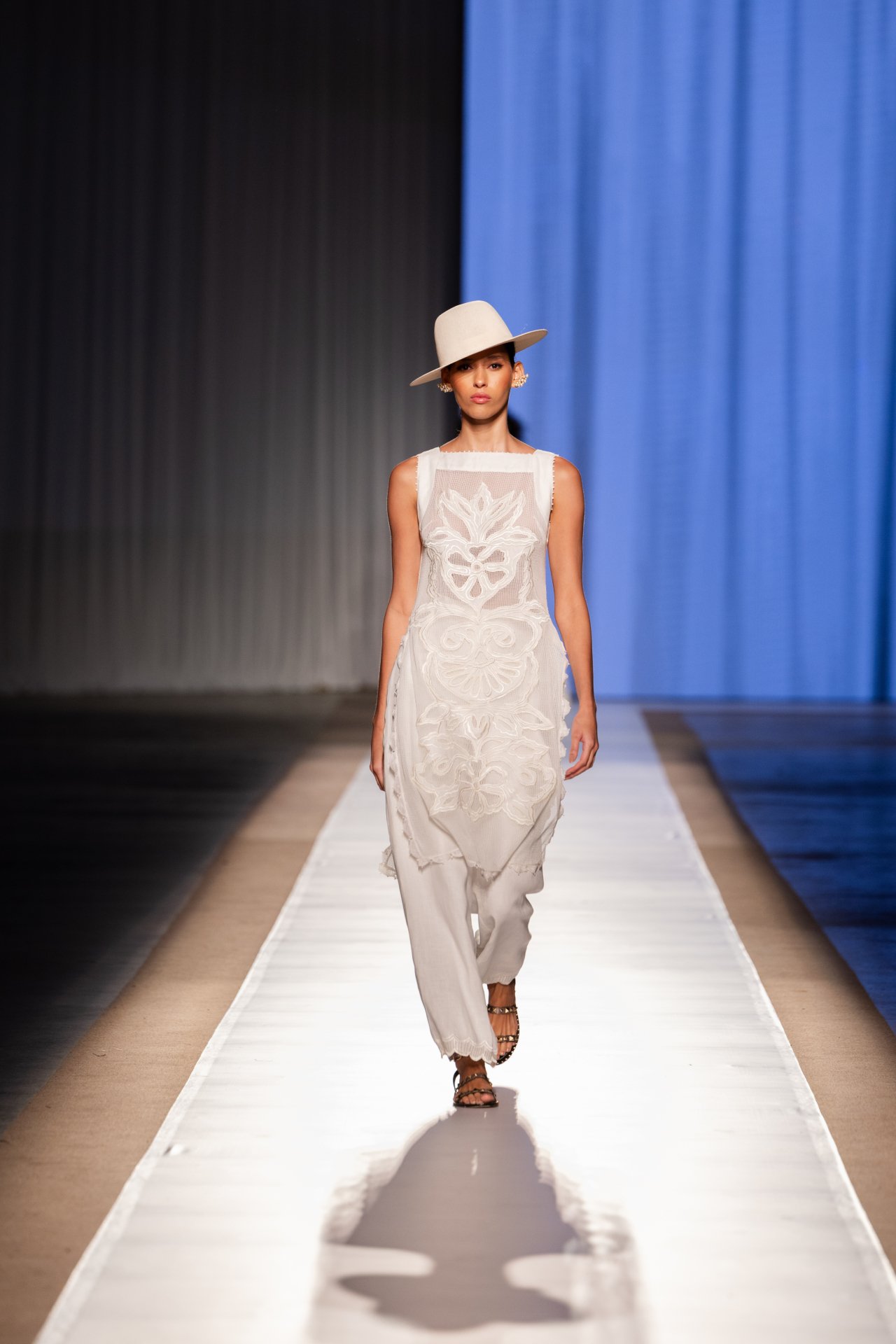Modelo em passarela com vestido branco rendado e chapéu bege no DFB Festival 2025.
