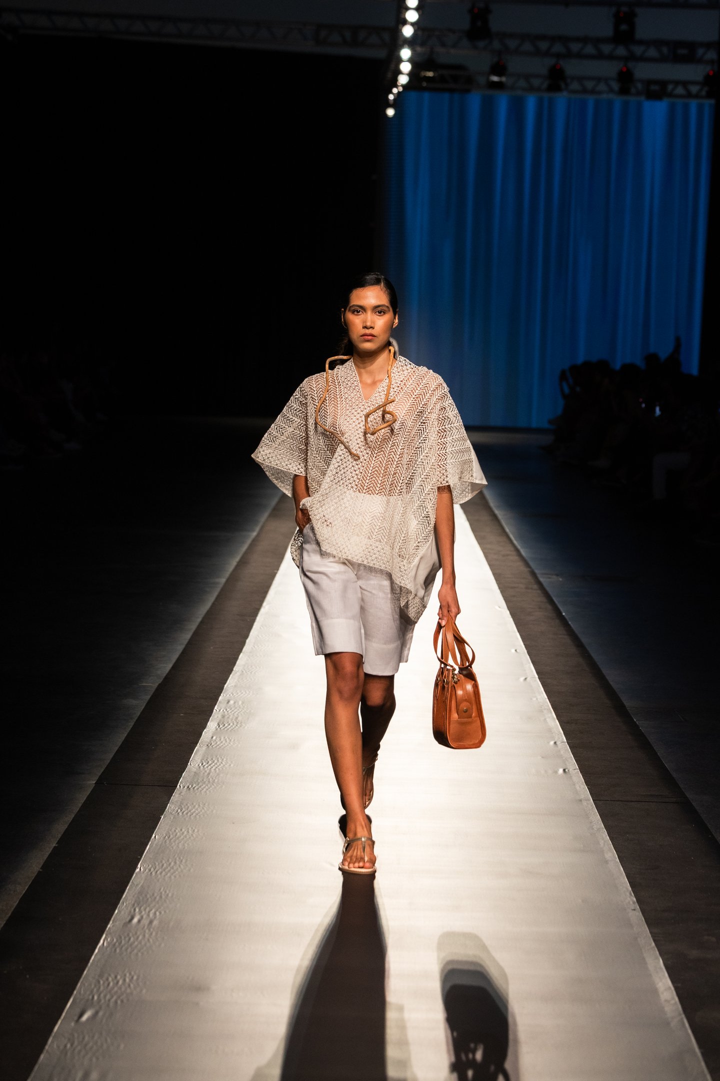 Modelo desfilando em passarela, usando roupa leve e bolsa de couro, durante o DFB Festival 2025.