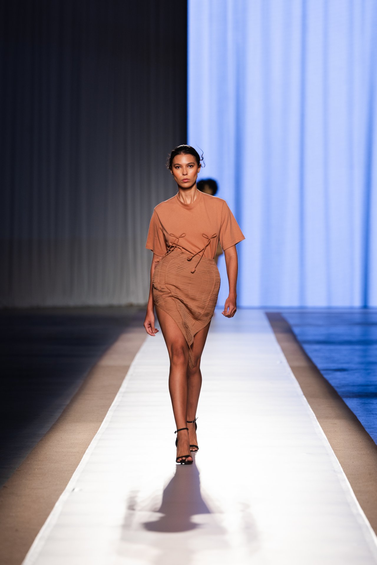 Modelo desfila com vestido marrom em passarela no DFB Festival 2025, fundo azul e cortina cinza.
