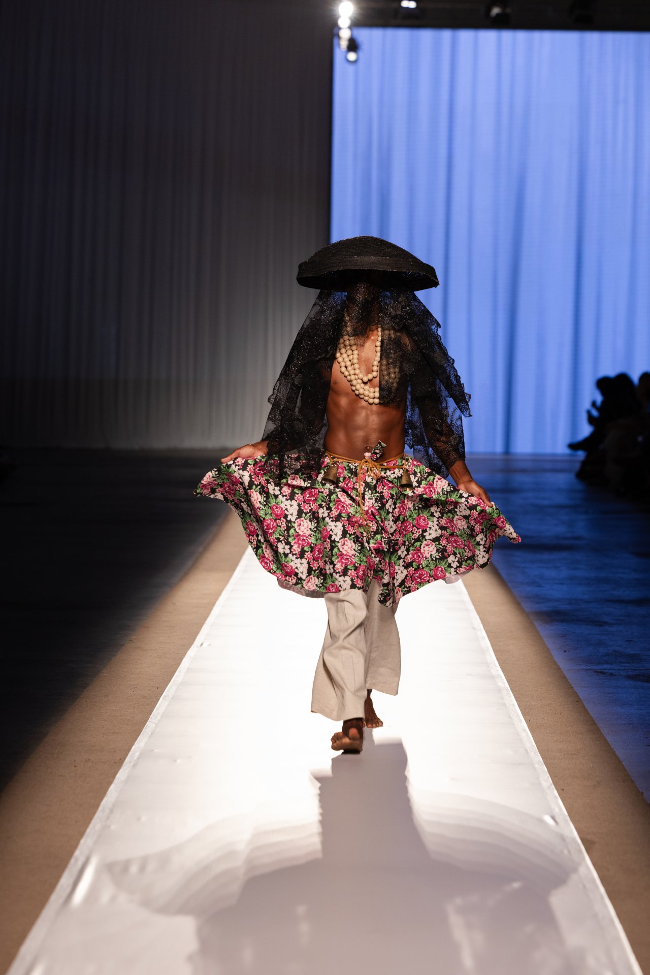 Modelo com chapéu e véu preto, saia floral e colar de pérolas na passarela do DFB Festival 2025.