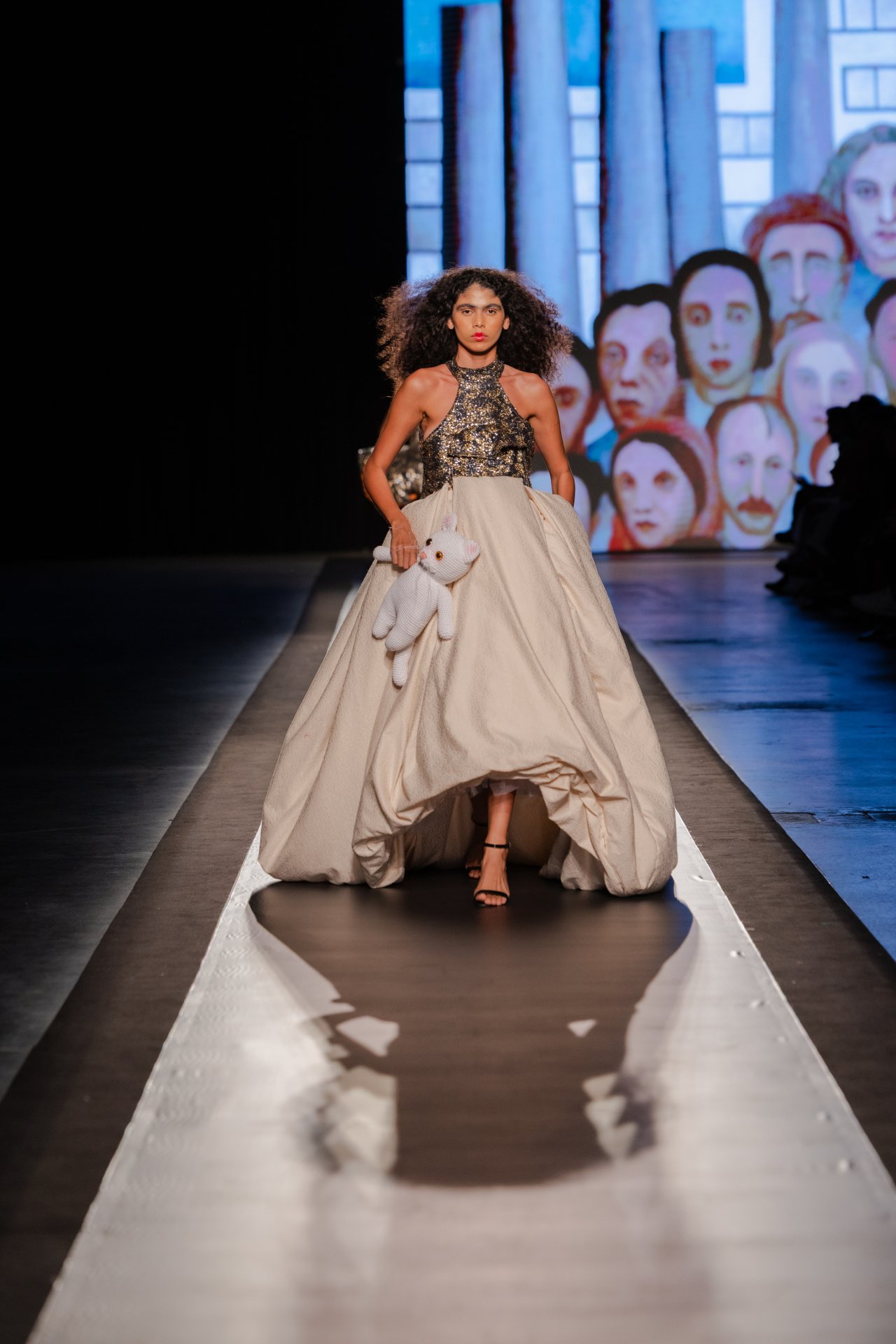 Modelo desfila em passarela com vestido glamoroso e urso de pelúcia no DFB Festival 2025. Ao fundo, arte colorida.