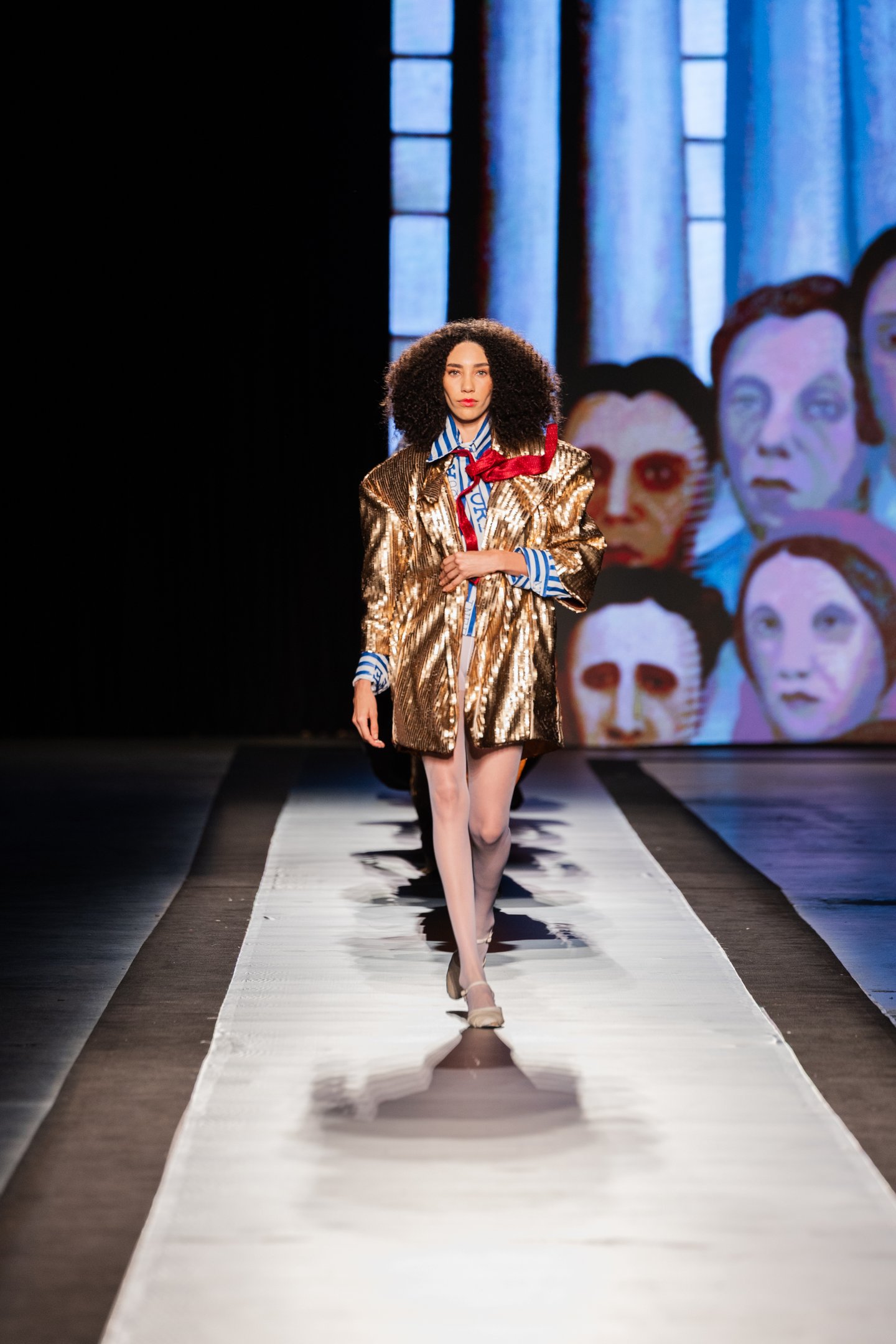 Modelo desfila com casaco dourado brilhante e camisa listrada no DFB Festival 2025; fundo com arte de rostos estilizados.