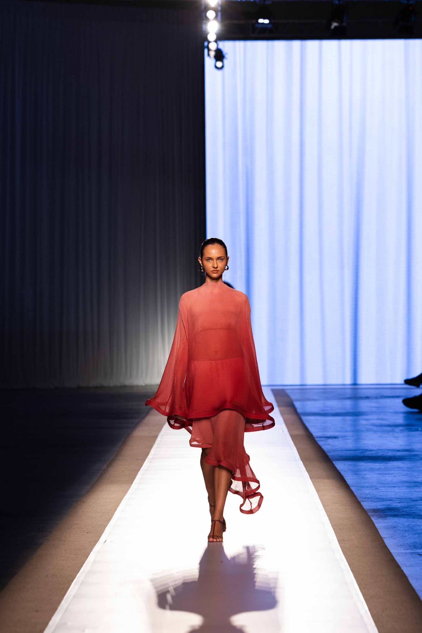 Modelo com vestido bege e óculos brancos desfilando, segurando bolsa vermelha no DFB Festival 2025.