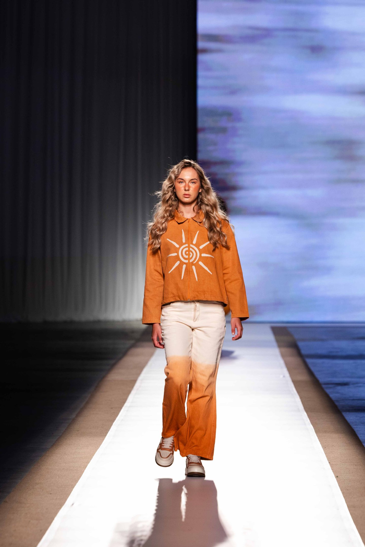 Modelo desfila com roupa em tons de laranja e bege em passarela do DFB Festival 2025.