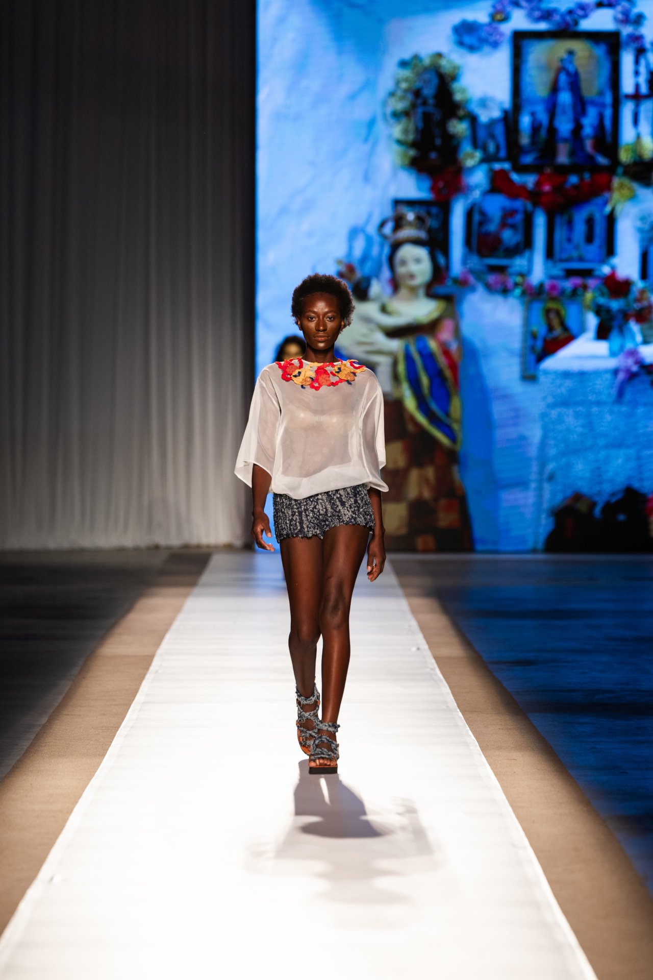 Modelo desfilando com blusa translúcida e detalhe floral no DFB Festival 2025. Ao fundo, arte colorida.