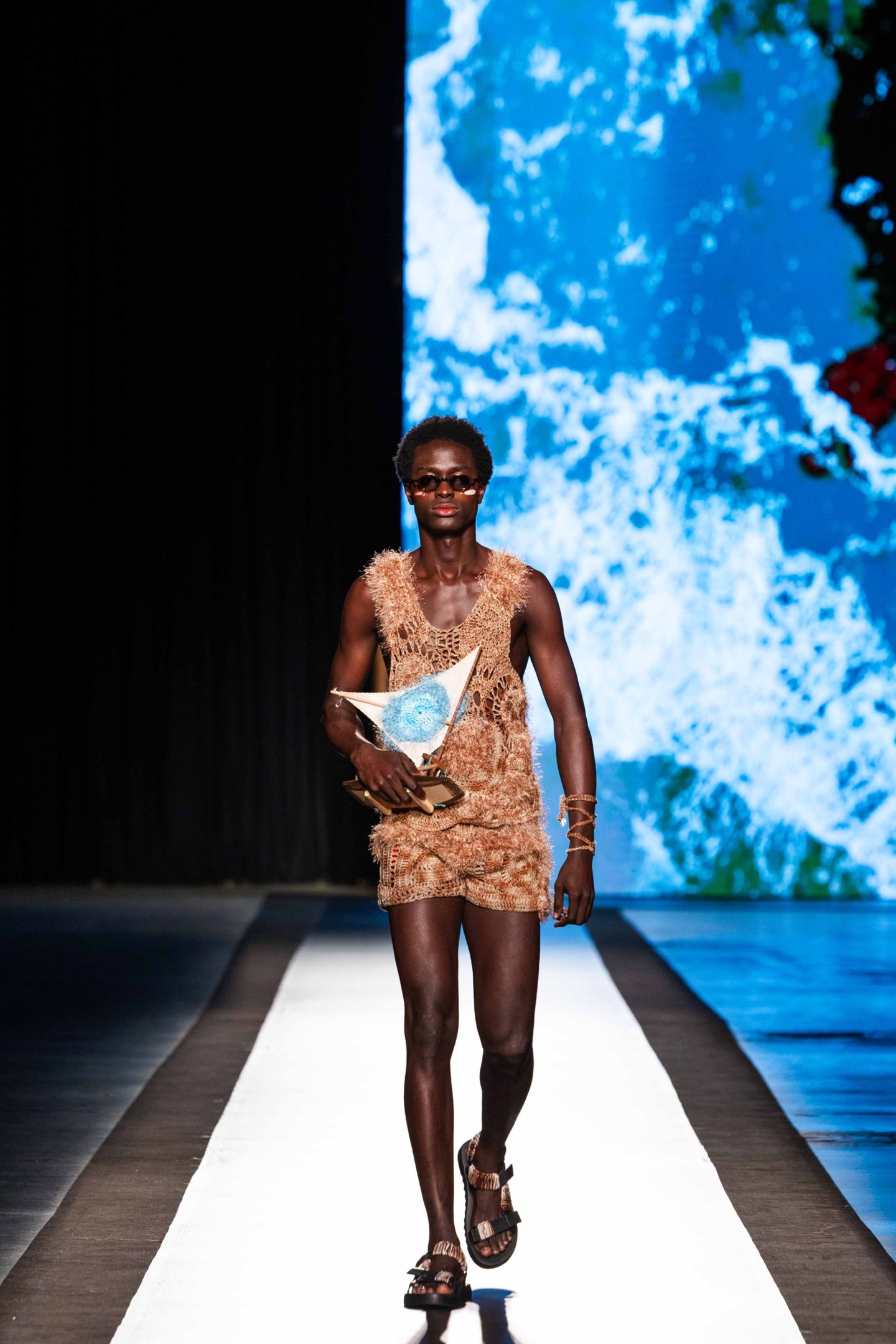 Modelo desfila com roupa texturizada e acessórios na passarela do DFB Festival 2025, fundo azul abstrato.