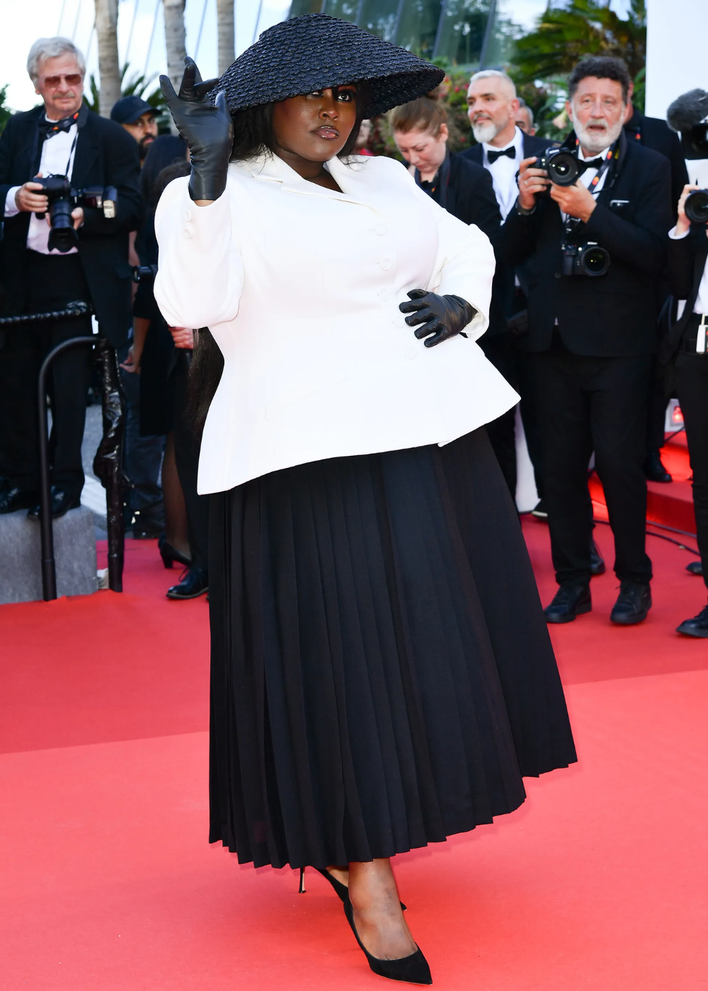 Pessoa usando chapéu preto e luvas, com terno branco e saia preta em evento. Fotógrafos ao fundo.
