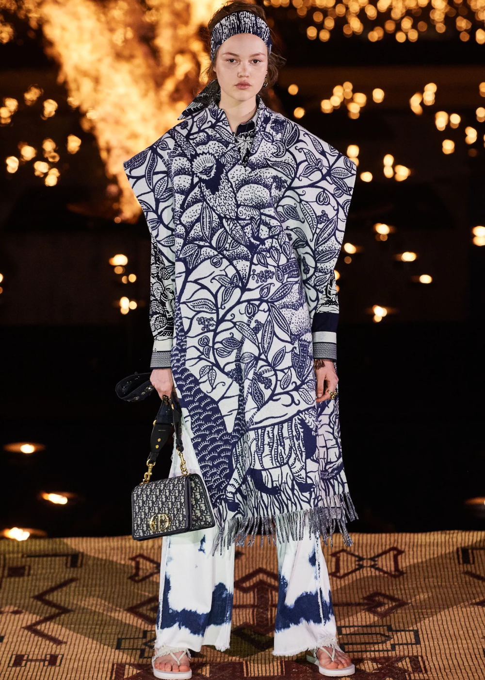 Vestido estampado azul e branco com padrões botânicos, bolsa Dior e cenário iluminado ao fundo.