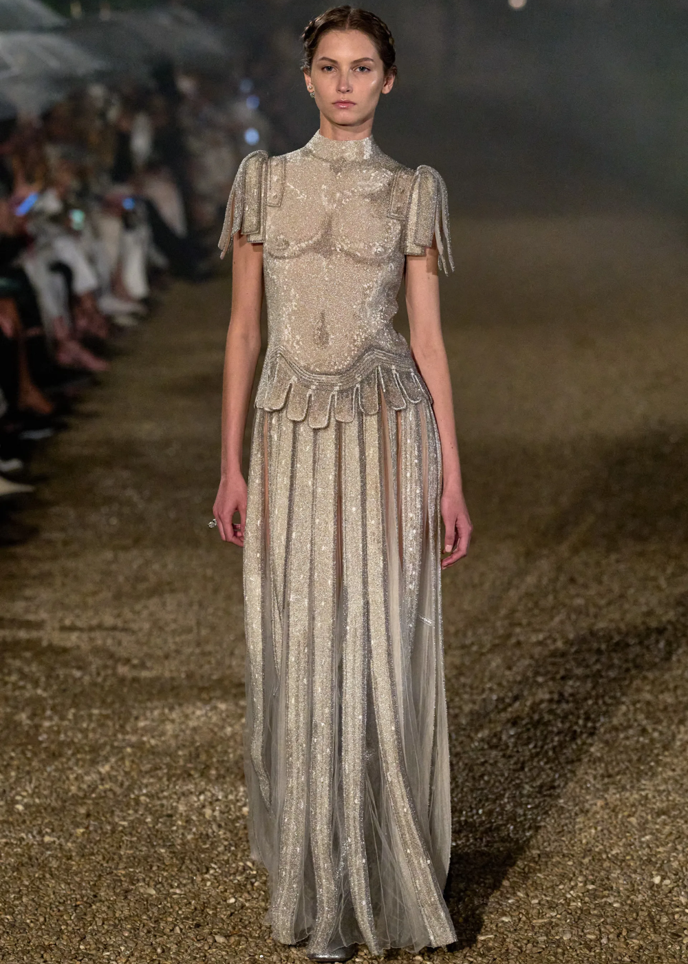 Modelo com vestido brilhoso e detalhes elegantes caminha em passarela durante desfile noturno. Ligação com Maria Grazia Chiuri.