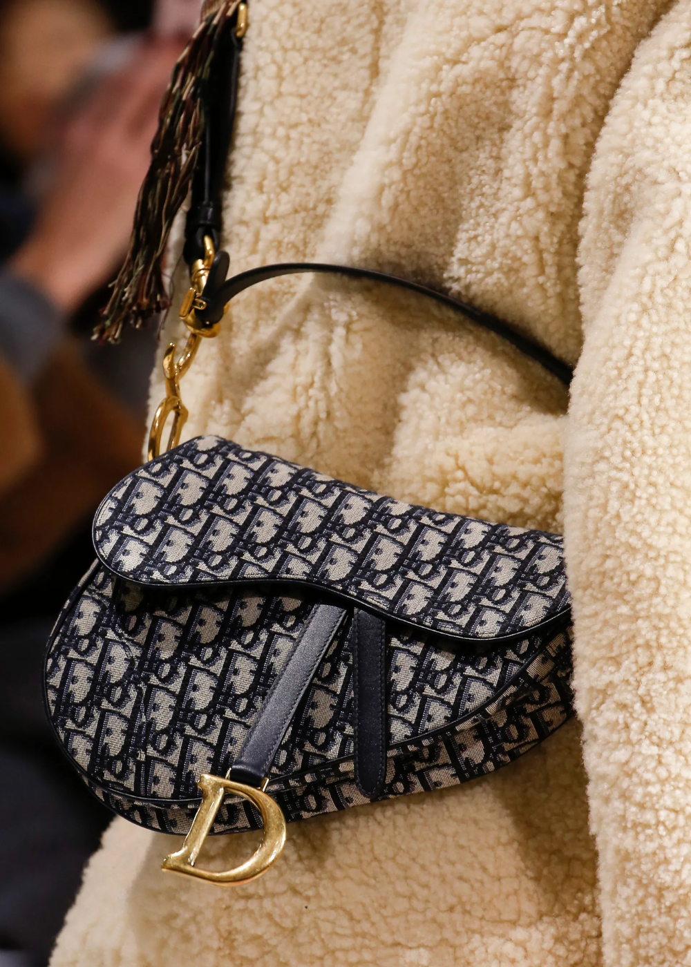 Bolsa de ombro Dior com padrão de monograma azul marinho e detalhes em metal dourado, sobre casaco de lã bege.