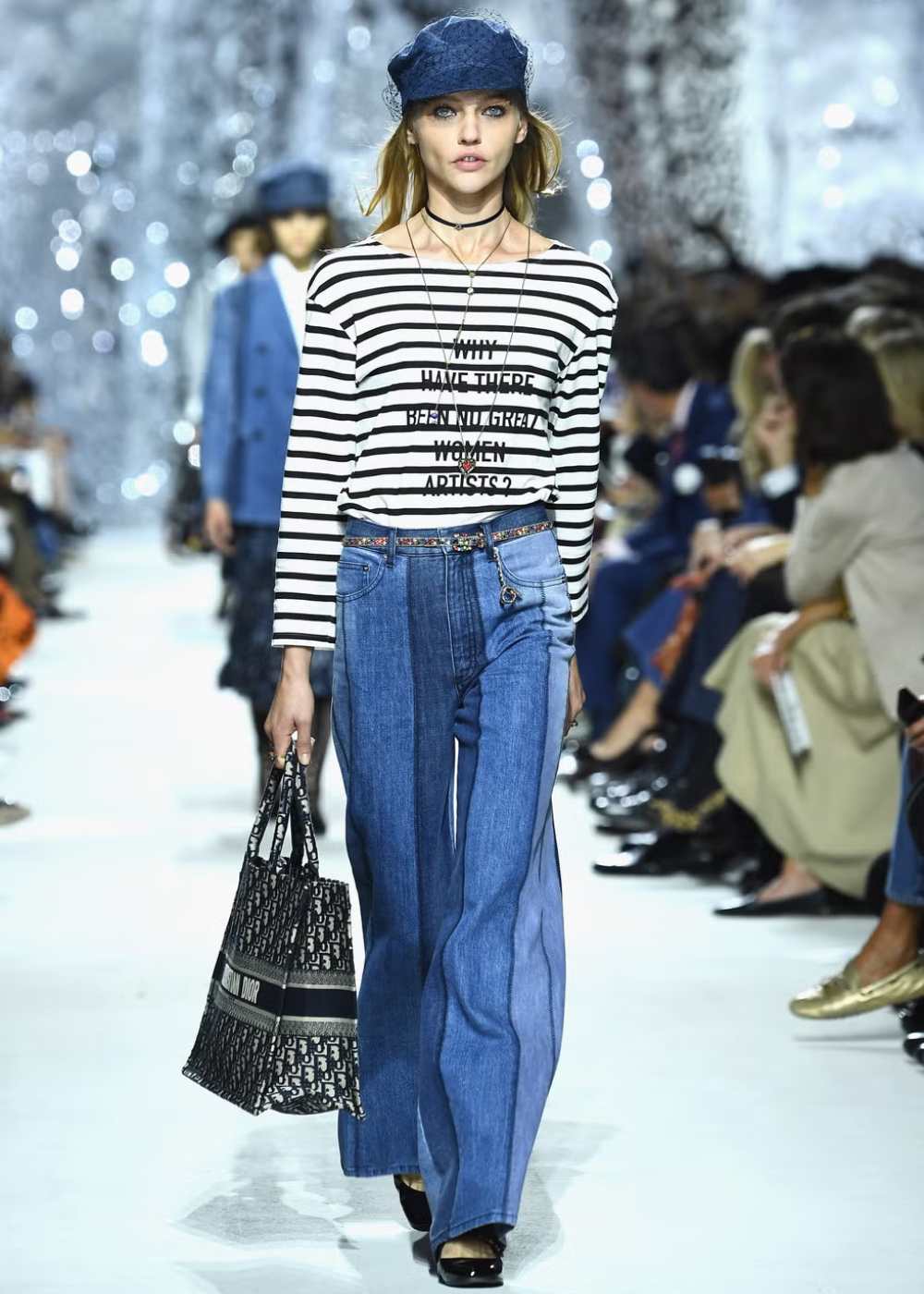 Desfile de moda com modelo vestindo blusa listrada e calça jeans larga, em visual inspirado por Maria Grazia Chiuri.