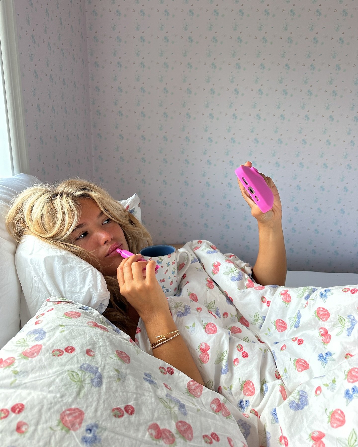 Loira deitada na cama, usando celular rosa e segurando caneca. Cenário de conforto, mas sugere distração.