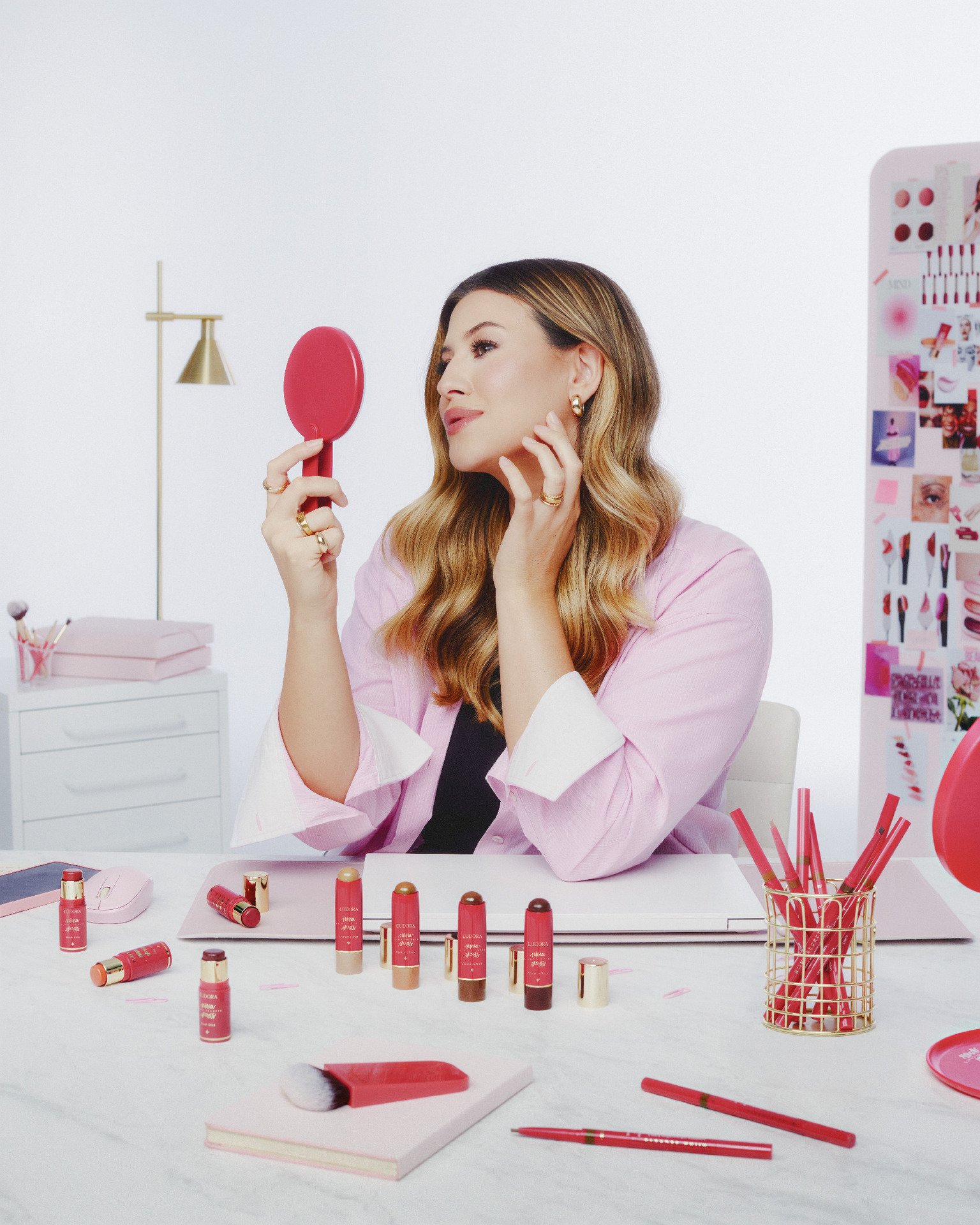 Mulher segura espelho em mesa com variados produtos em stick, itens de maquiagem e utensílios, em um ambiente claro e organizado.