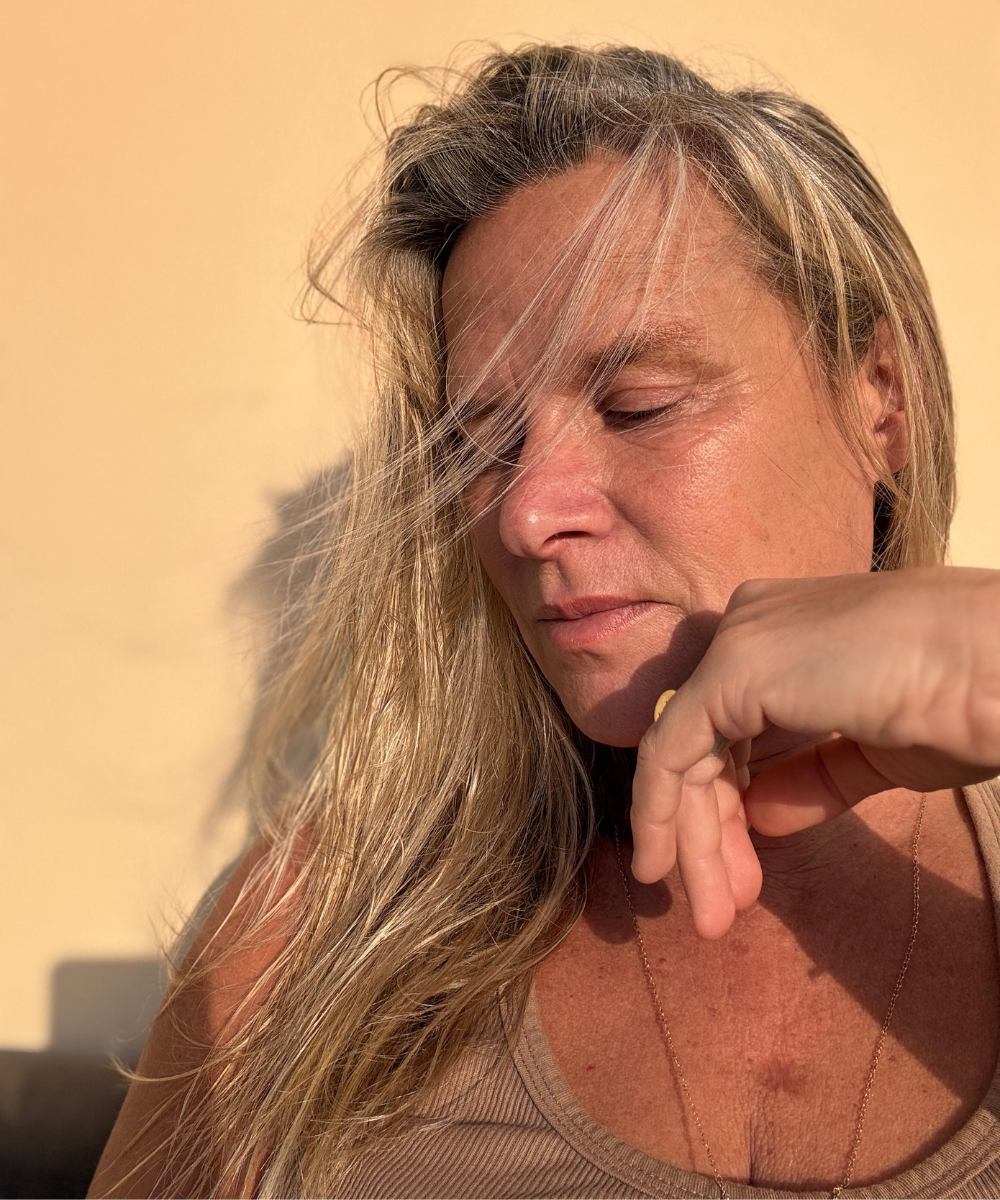 Mari Becker em close com expressão contemplativa, cabelos loiros ao vento, sob a luz suave do sol.