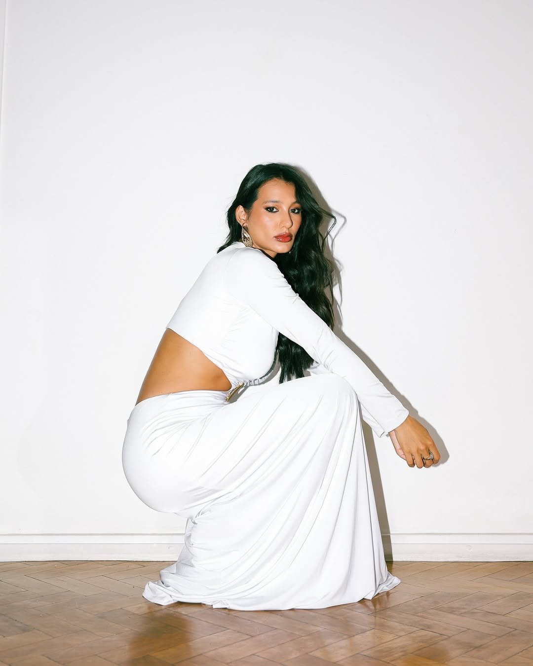 Mulher com vestido branco posando agachada em um fundo claro.