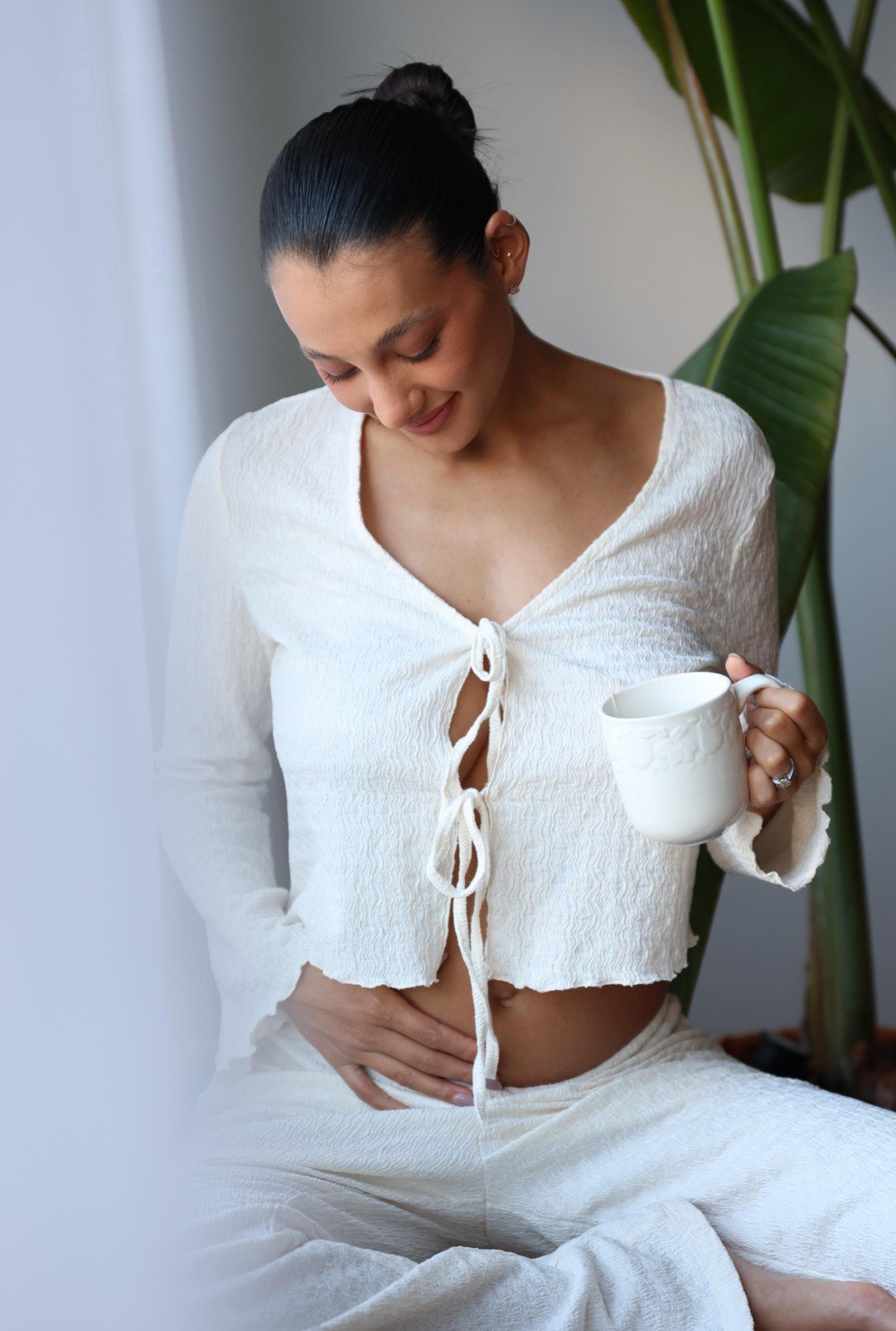 Mulher grávida sentada com uma xícara à mão, sorrindo suavemente e tocando a barriga.