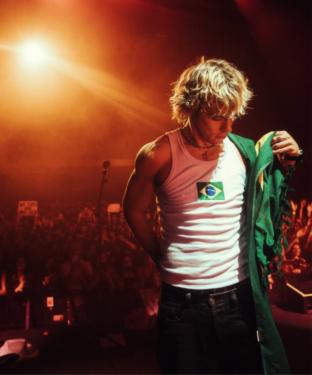 Jovem no palco com bandeira do Brasil e camiseta. Atmosfera de show vibrante. The Driver Era.