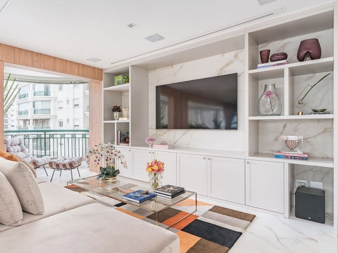 Sala moderna com estante na decoração ao redor da TV, sofás claros e mesa de centro com flores.
