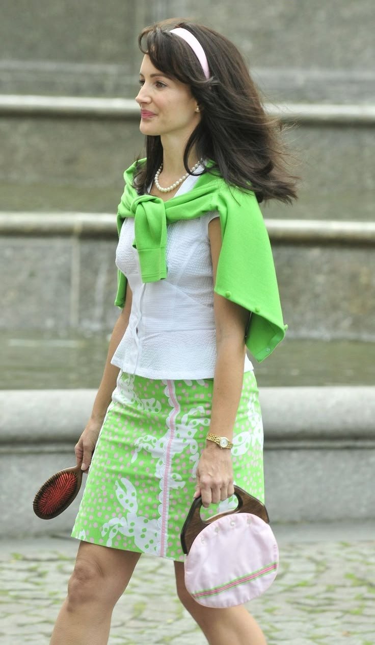 Mulher elegante com blusa branca, suéter verde sobre os ombros, saia estampada e bolsa, evocando o charme clássico.