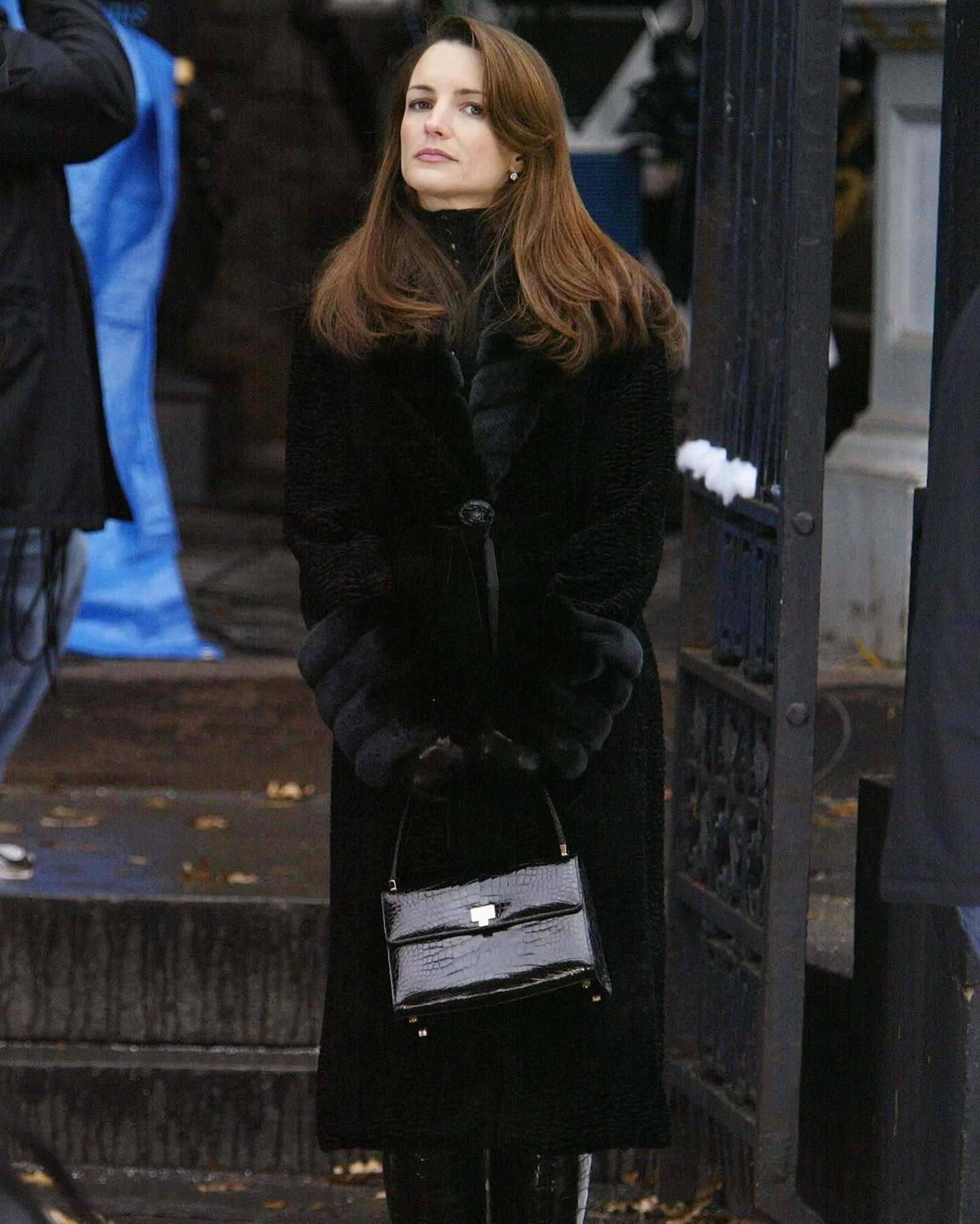 Pessoa com casaco elegante e bolsa de mão preta, refletindo o estilo clássico e sofisticado de Charlotte York.