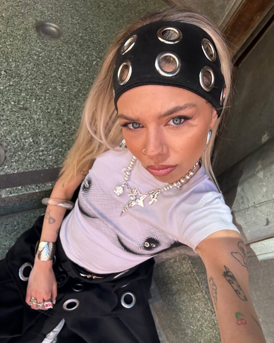 Pessoa com faixa de cabelo tendência decorada com ilhós metálicos. Usa acessórios prateados e camiseta com estampa ousada.