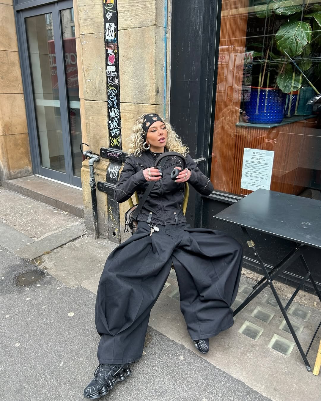Pessoa com faixa de cabelo tendência, usando calça larga preta senta em uma cadeira na rua.