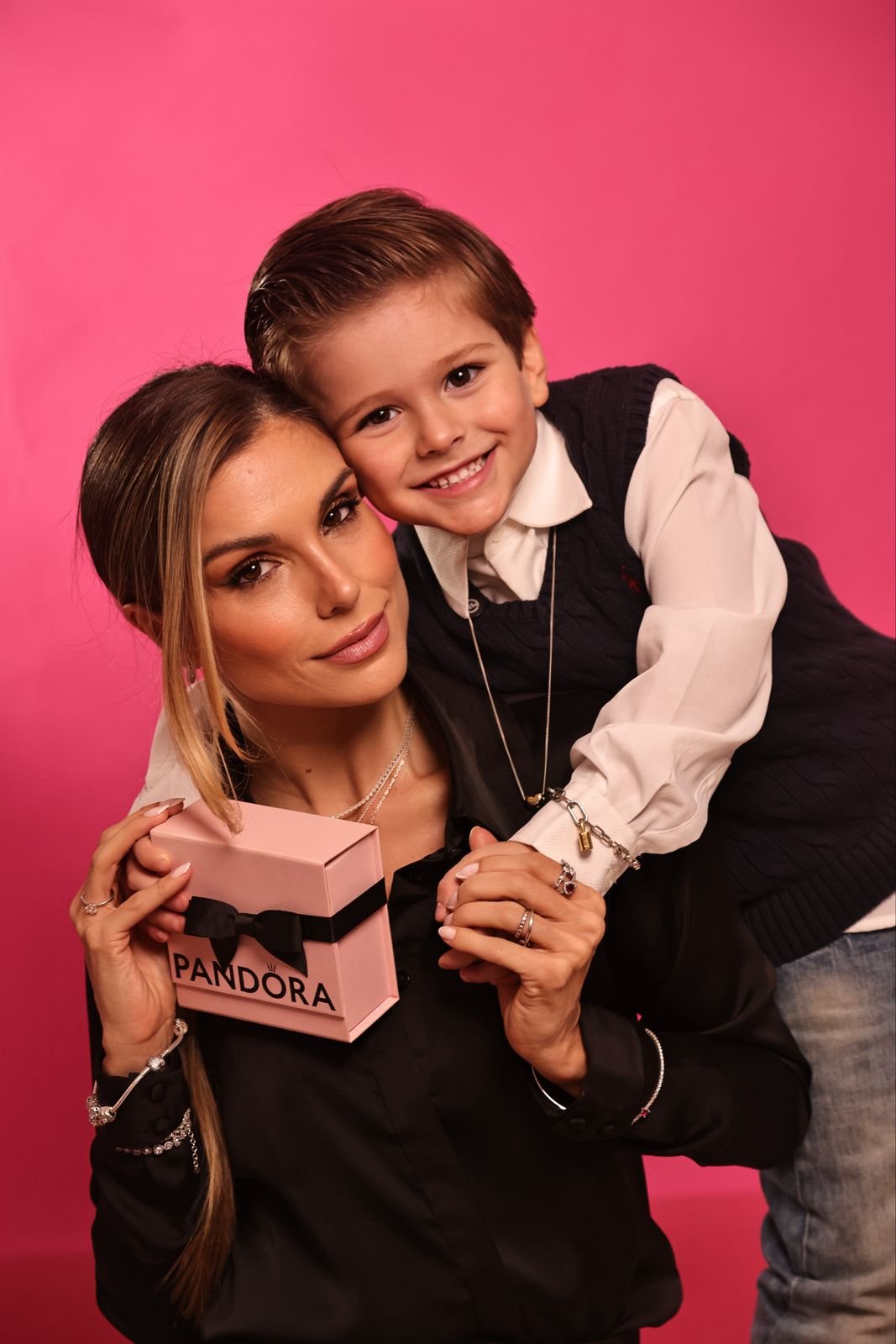 Flávia Viana segurando uma caixa da Pandora com uma criança sorrindo sobre seus ombros, contra um fundo rosa.