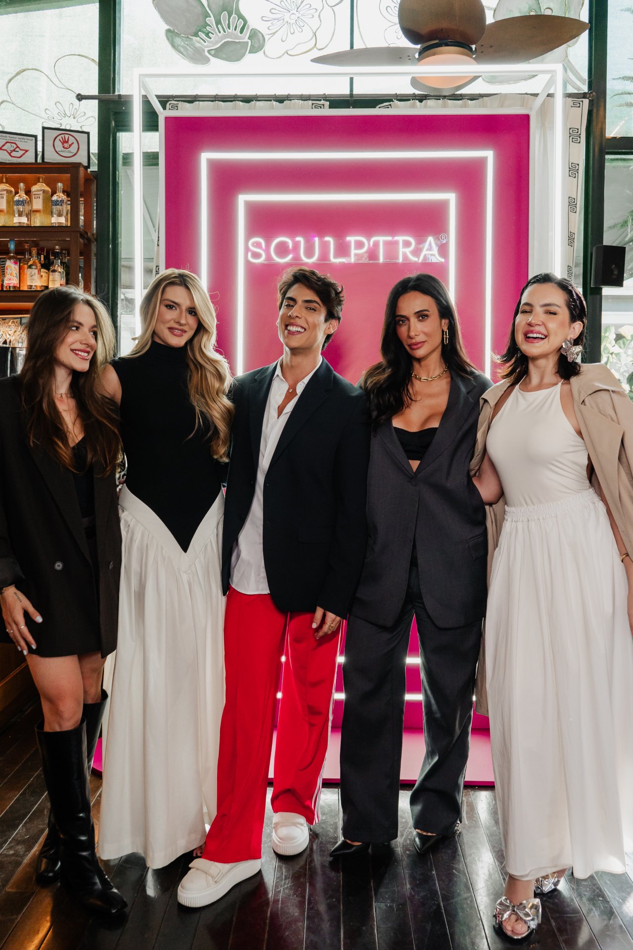 Pessoas posam sorrindo em frente a um painel rosa do evento Sculptra Experience.