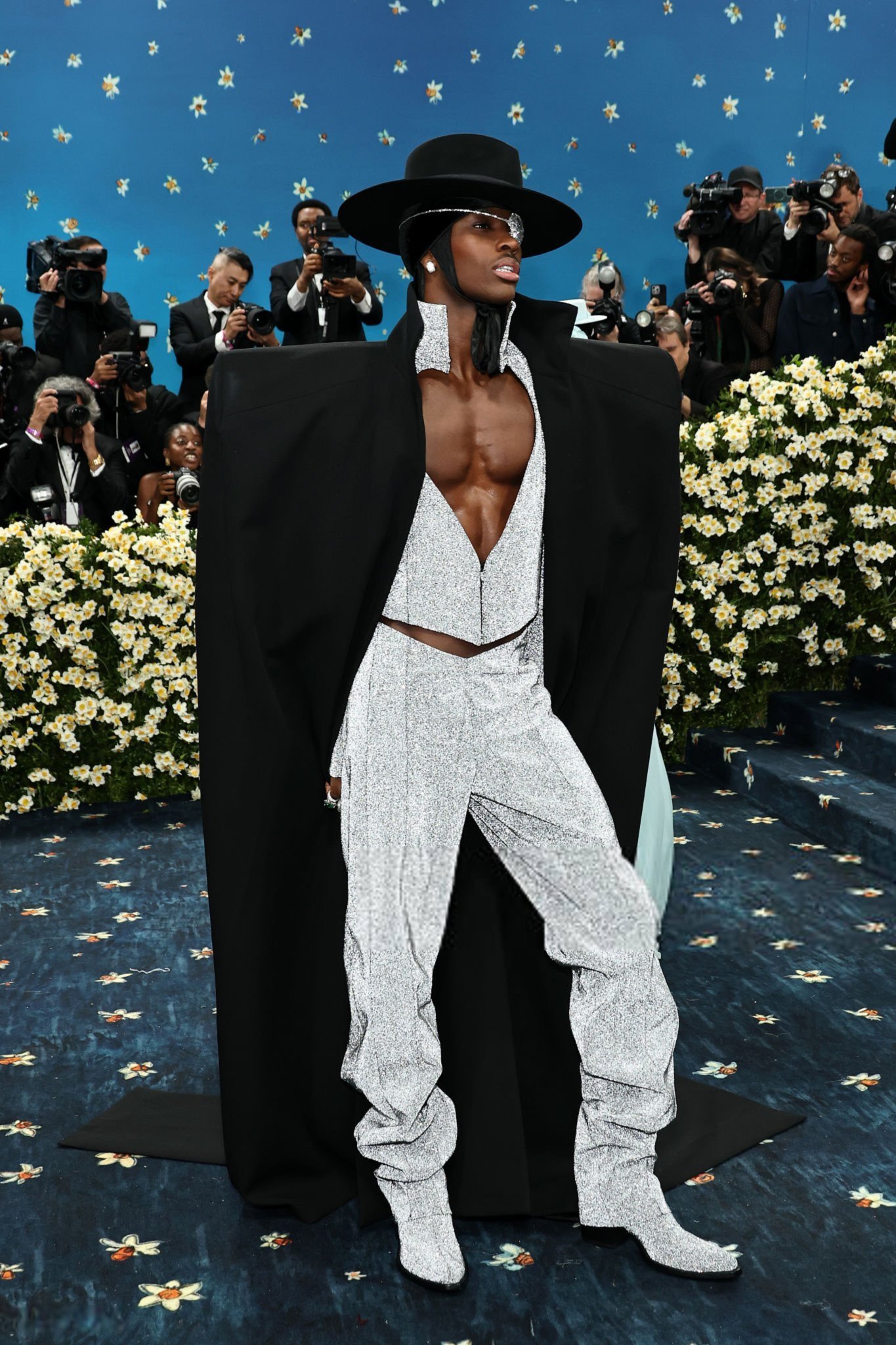 Homem de traje branco brilhante, capa preta e chapéu largo em evento formal, cercado por fotógrafos. looks masculinos