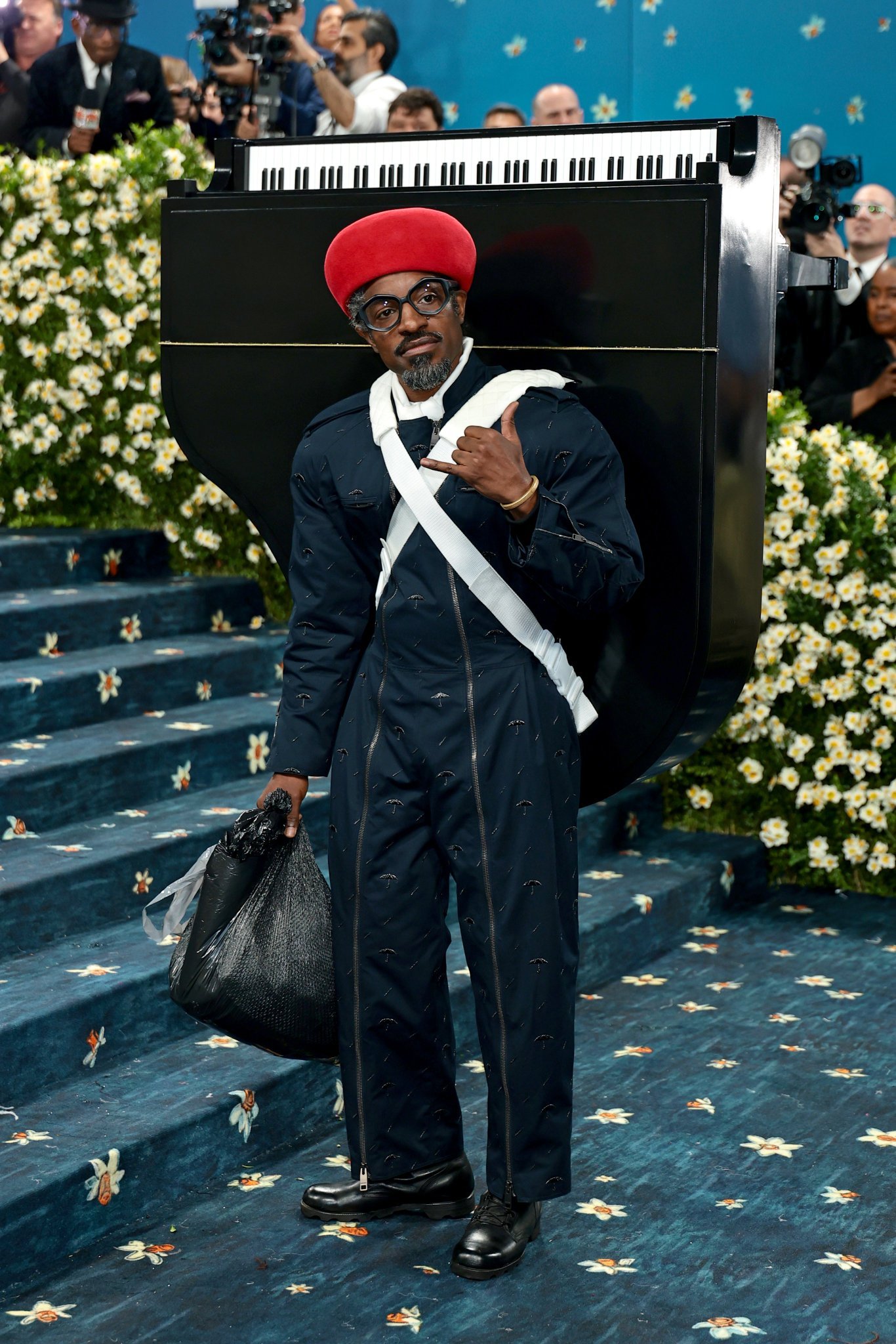 Um homem usa macacão azul marinho com chapéu vermelho, segurando uma bolsa, destacando looks masculinos criativos. Looks masculinos