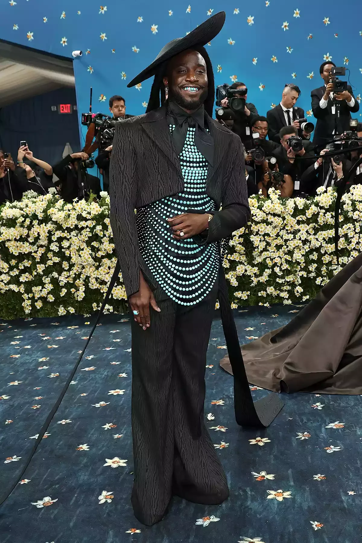 Celebridade com traje elegante preto, ornado com padrões verde-água e chapéu estiloso, em tapete vermelho. Looks masculinos