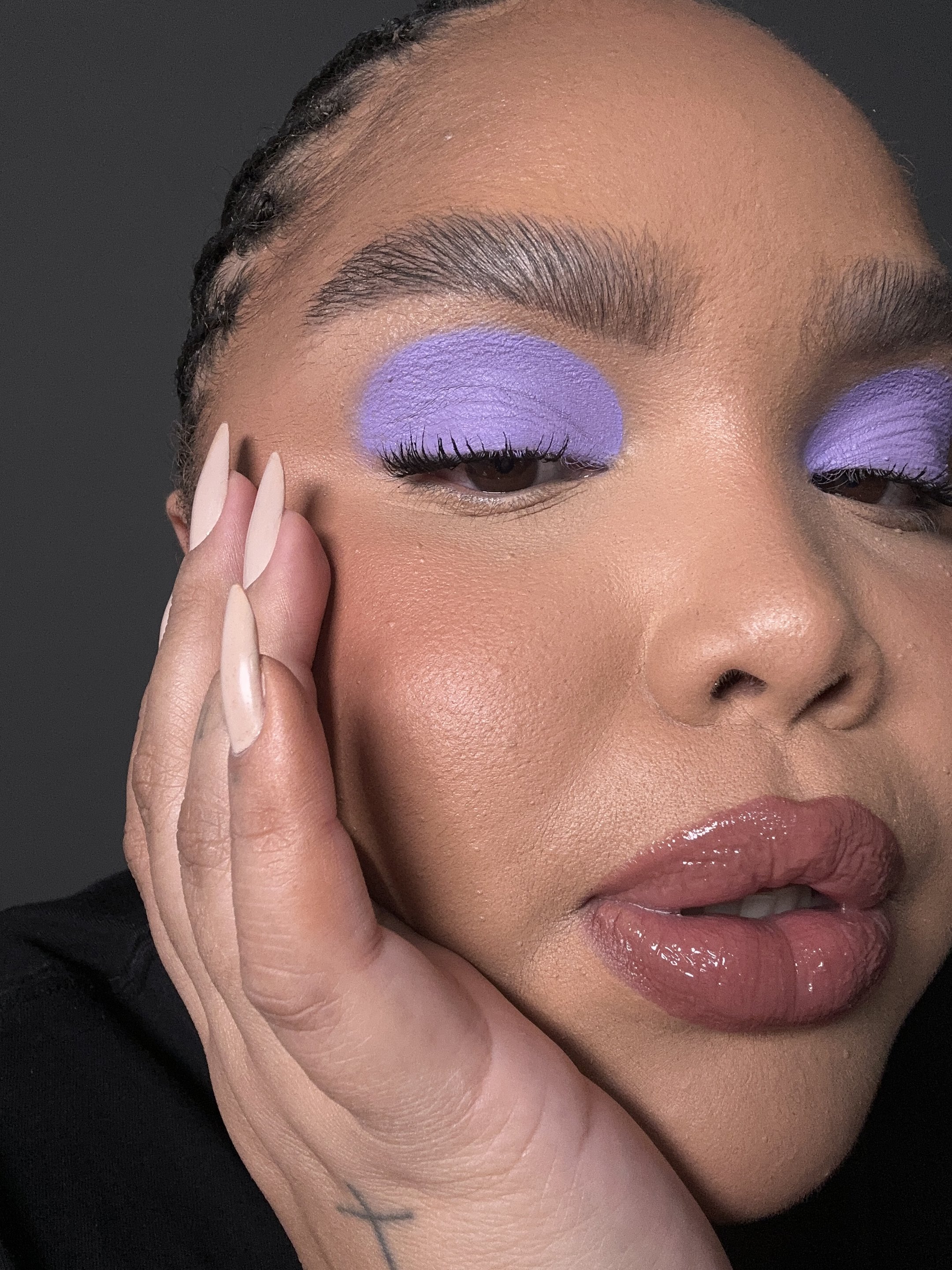 Sombra roxa destaca os olhos em maquiagem elegante; mão com unhas longas apoia o rosto.