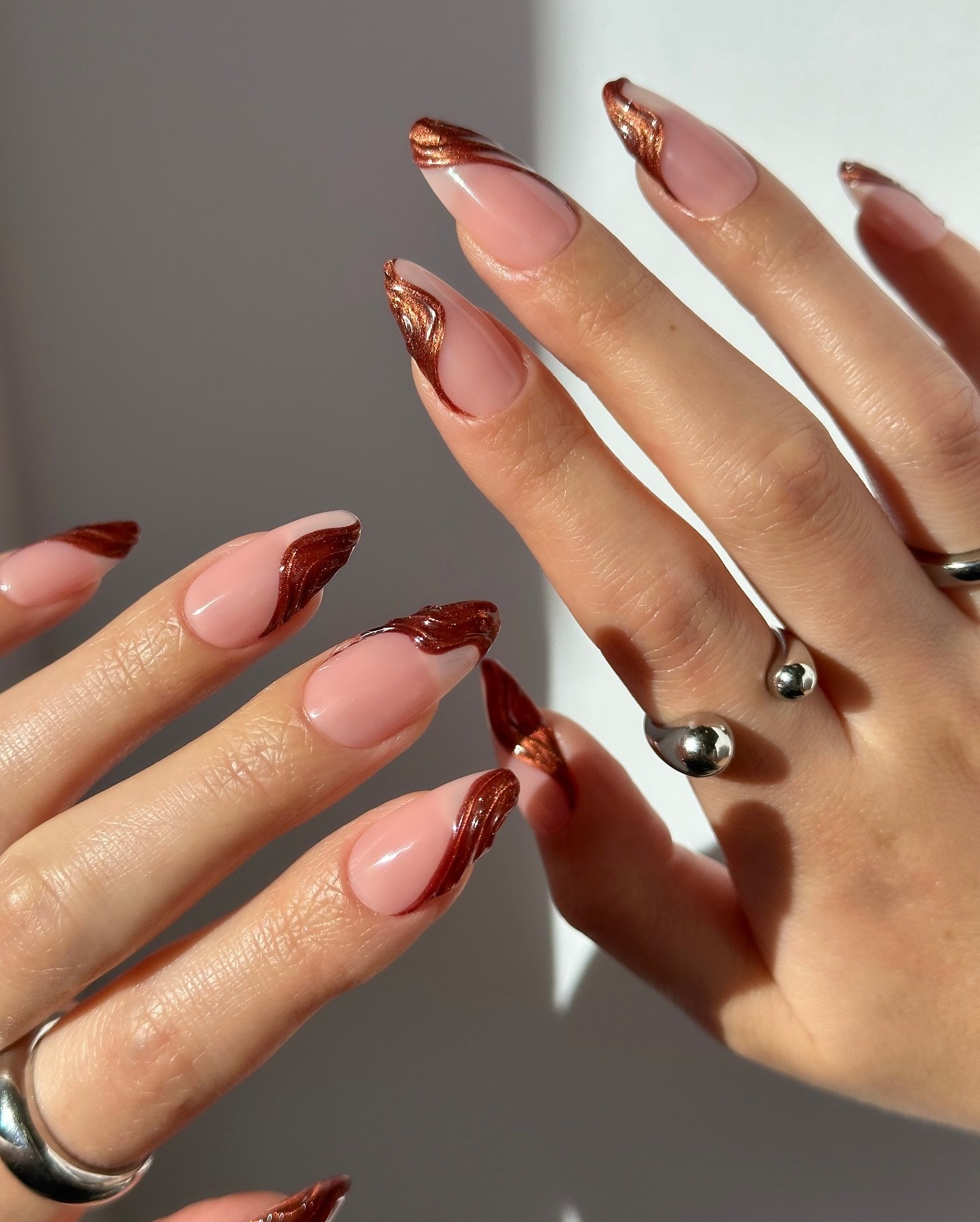 Unhas com esmalte nude e detalhes em espiral marrom metálico. Mãos com anéis prateados.