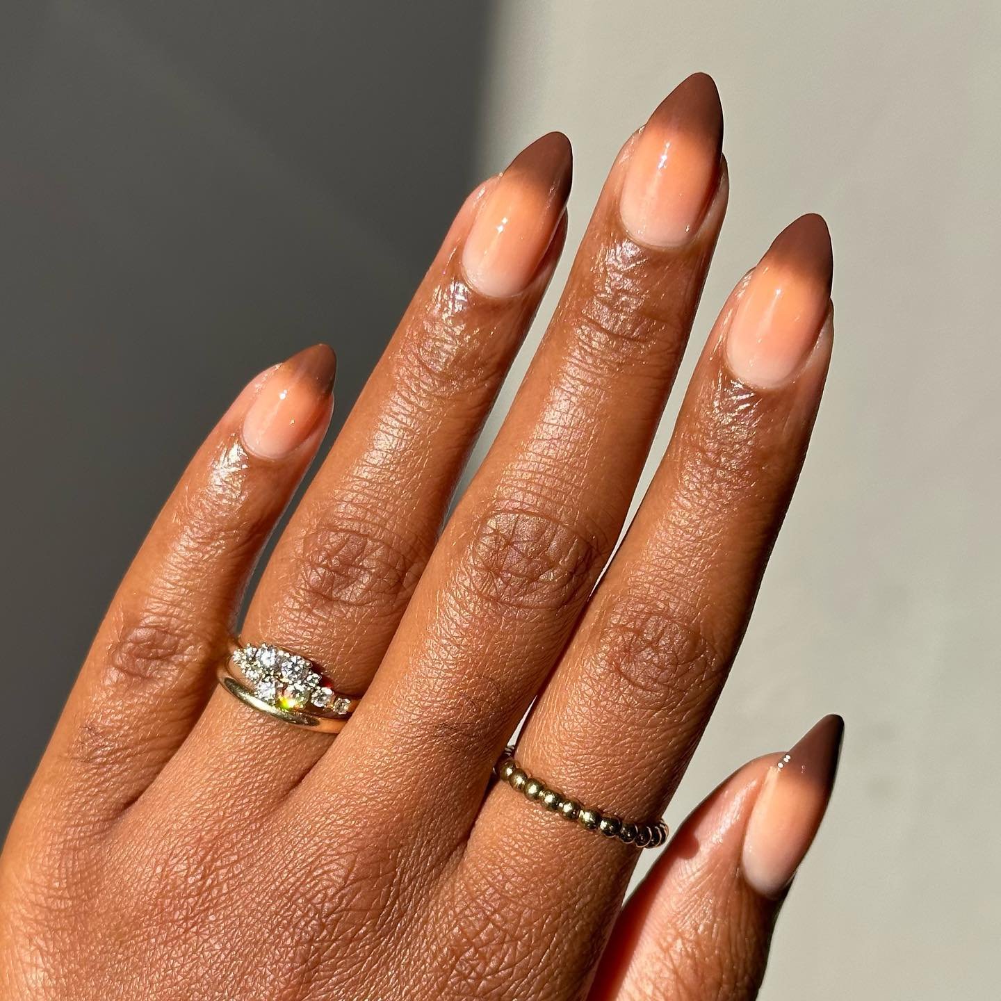 Mão com unhas marrons em degradê, joias douradas e anel brilhante.