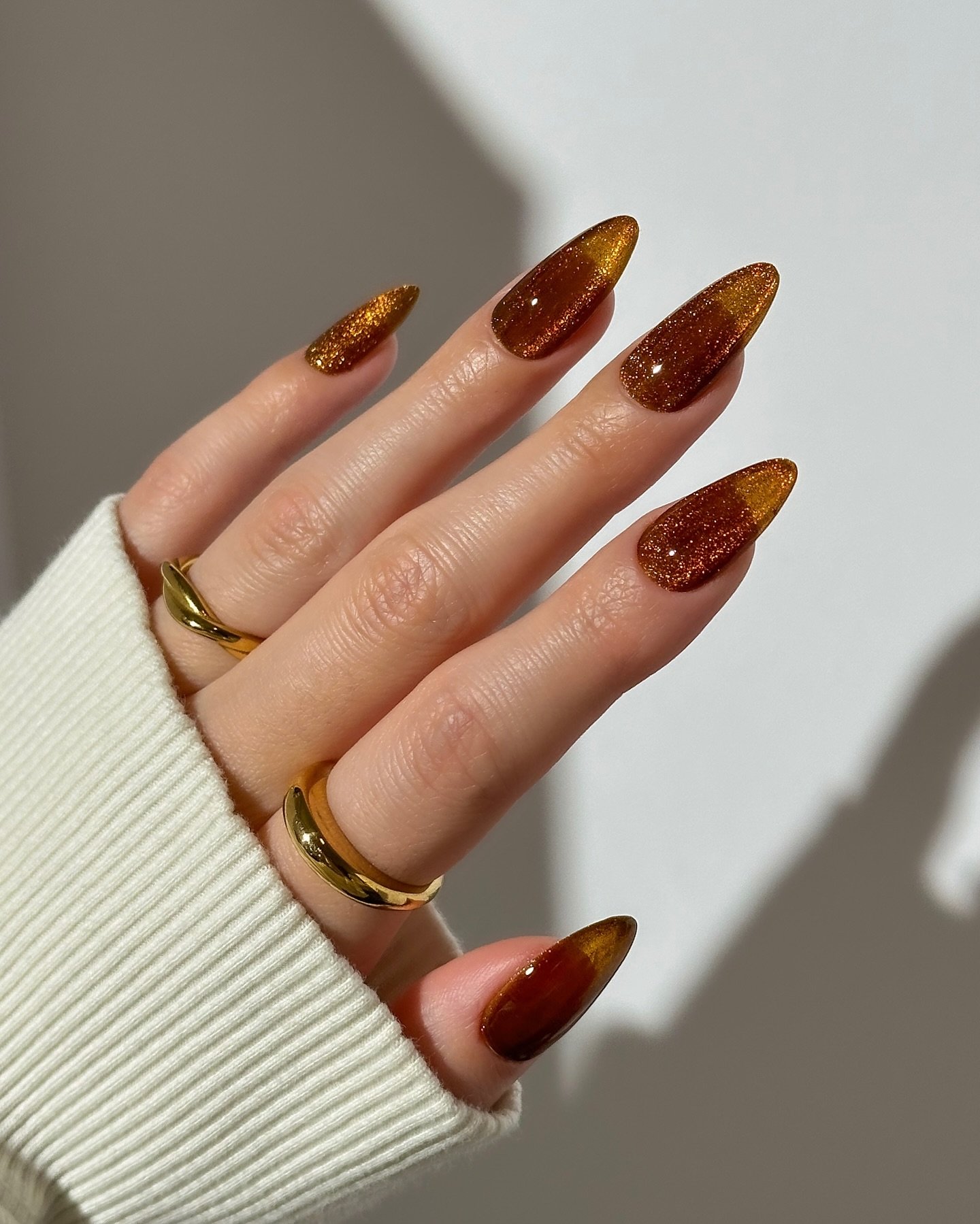Unhas marrom brilhante em formato amendoado, combinadas com anéis dourados.