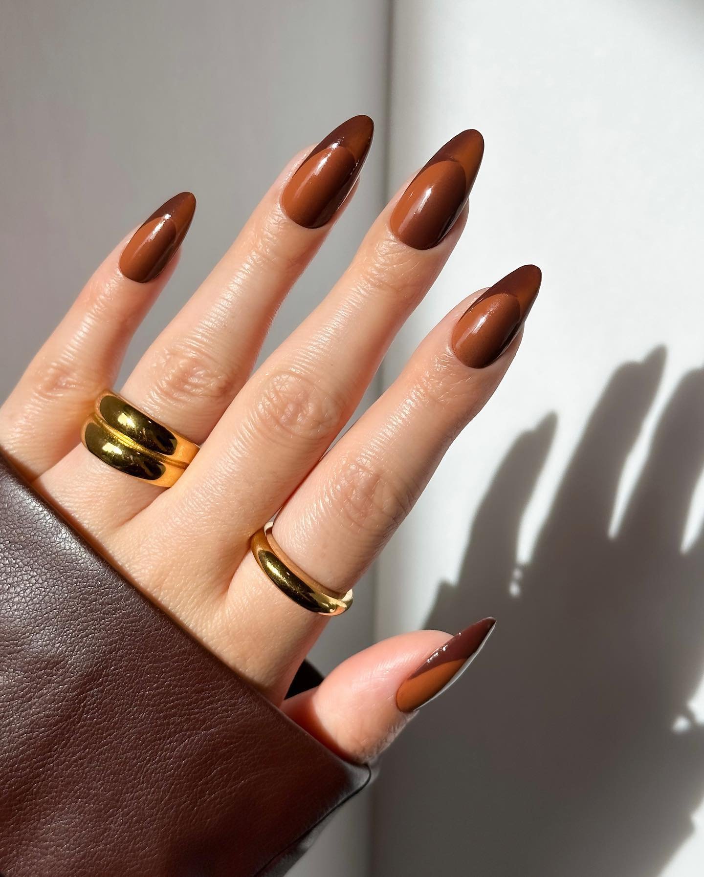 Unhas marrons em formato amendoado, mãos com anéis dourados e jaqueta marrom.