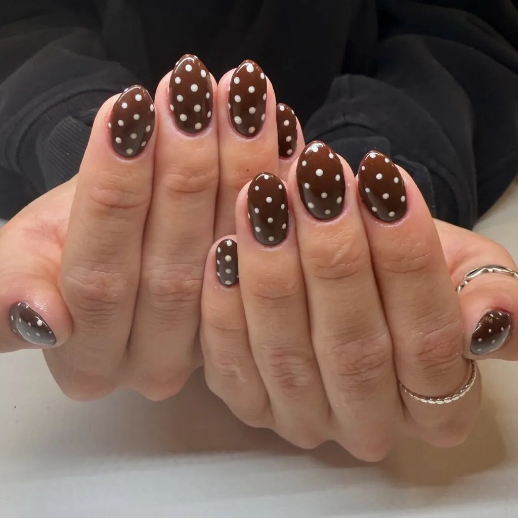 Unhas marrons decoradas com bolinhas brancas, exibidas em duas mãos juntas.
