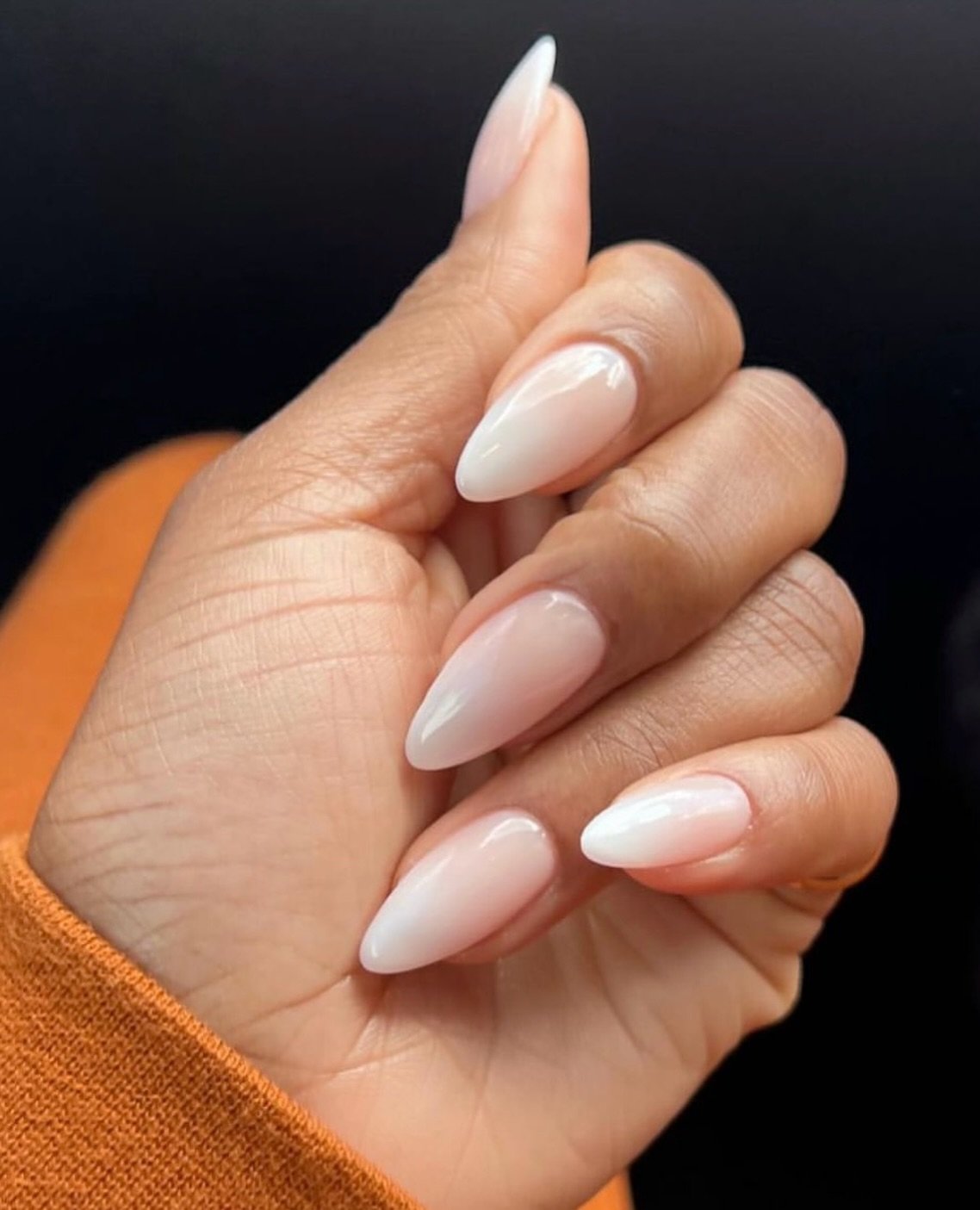 Unhas de gel simples e elegantes em formato amendoado e tom nude, destacam-se sobre fundo laranja.
