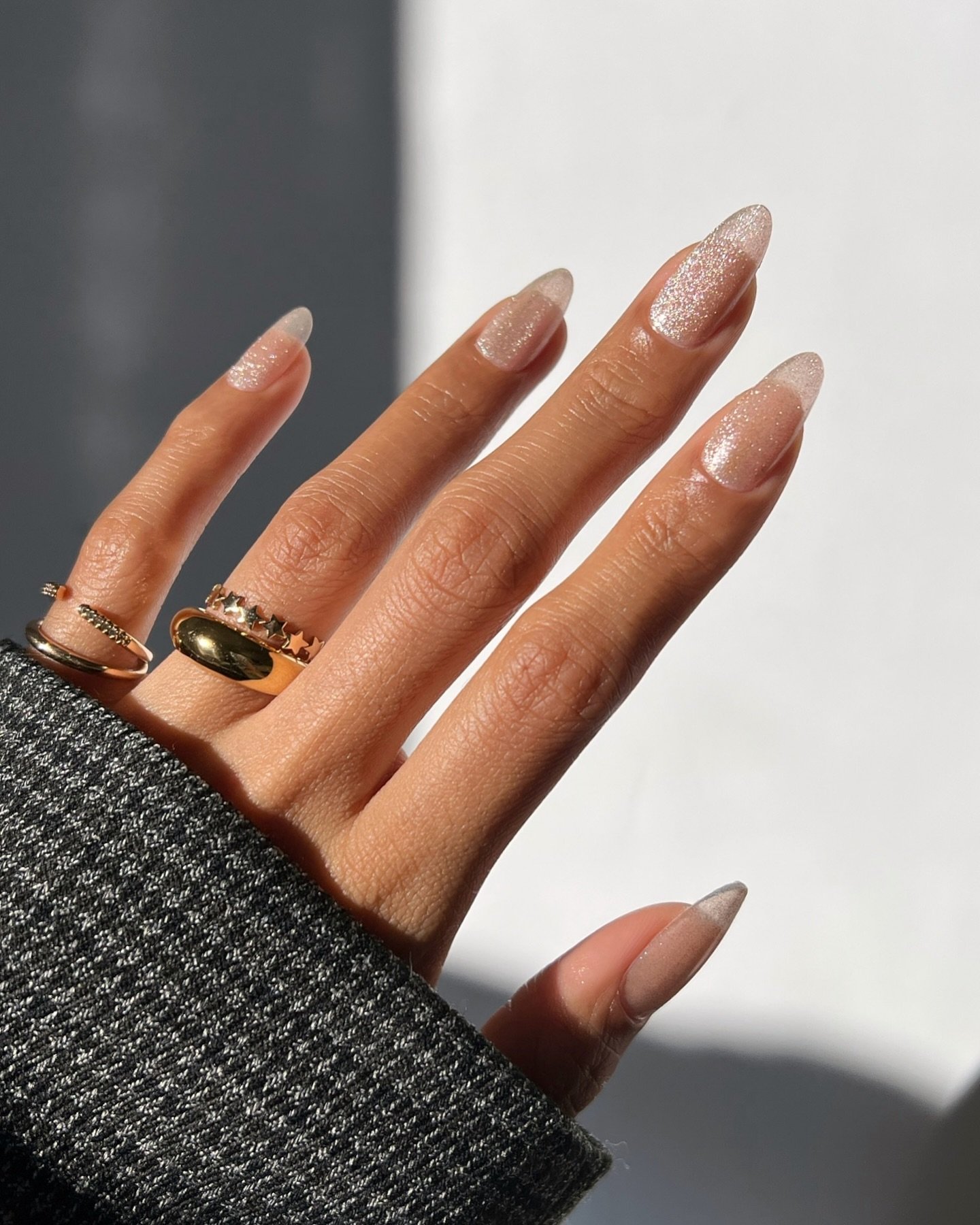 Mão com unhas de gel simples e elegantes, decoradas com brilho sutil, usando anéis prateados e dourados.