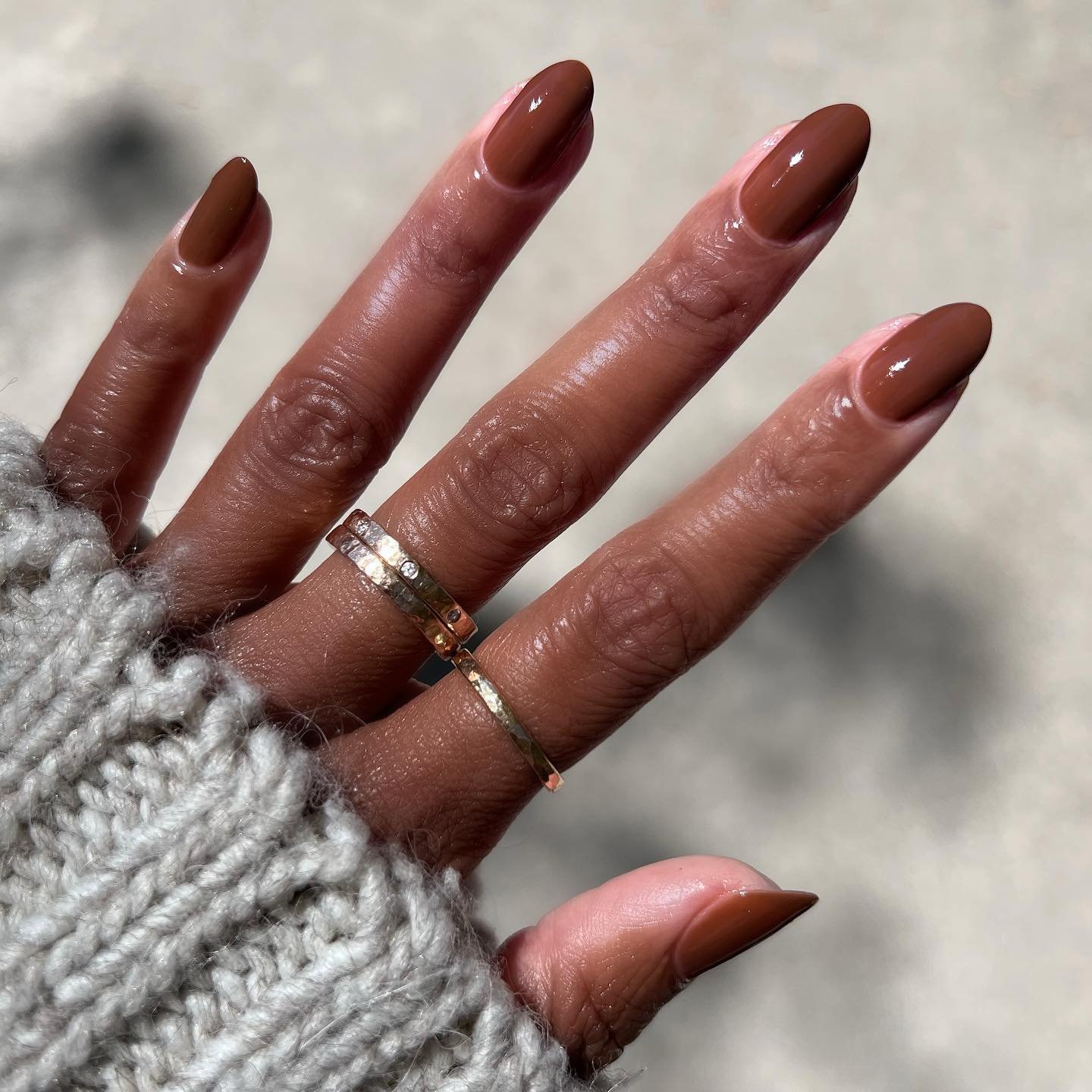 Unhas de gel marrons, simples e elegantes, com anéis dourados em mão com suéter cinza.