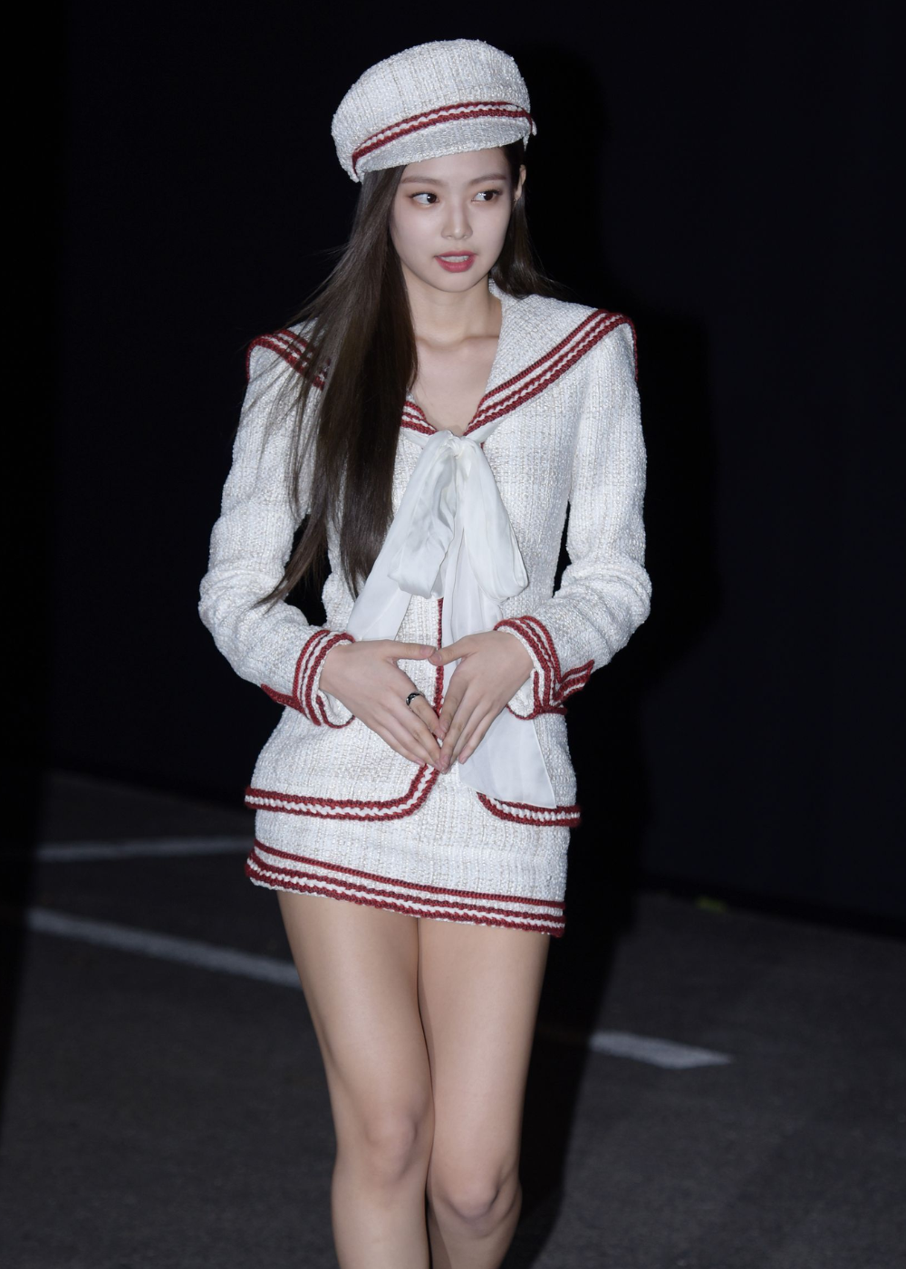 Jennie usando conjunto branco com detalhes vermelhos e chapéu, com expressão atenta.