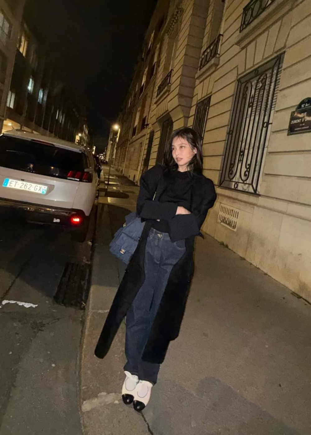 Mulher de preto em rua à noite, ao lado de carro branco estacionado. Jennie veste calça escura e sapatos claros.