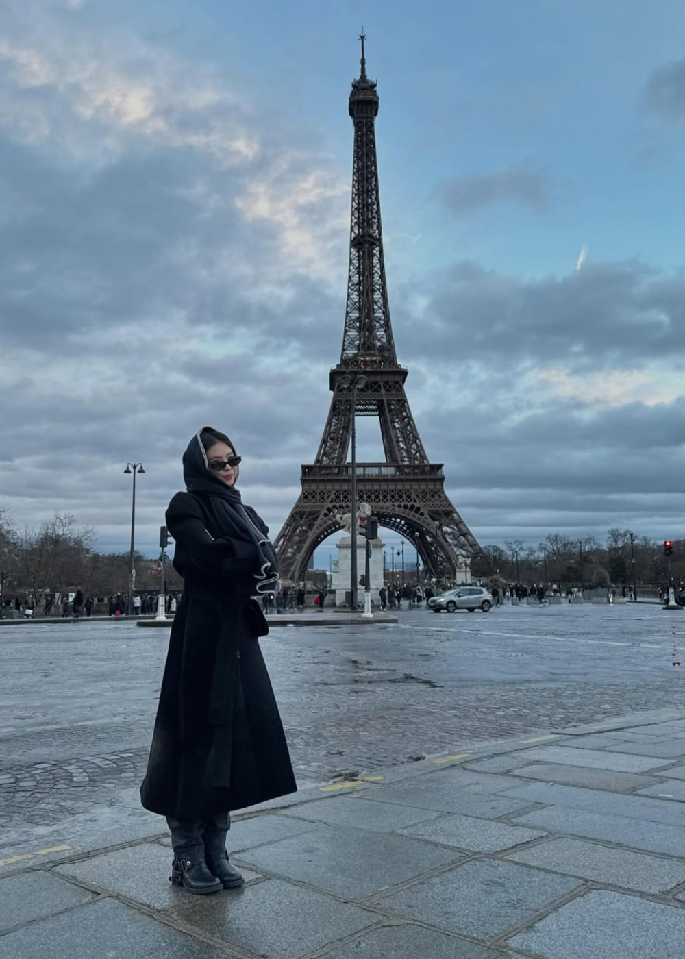 Pessoa de casaco preto em frente à Torre Eiffel, céu nublado.